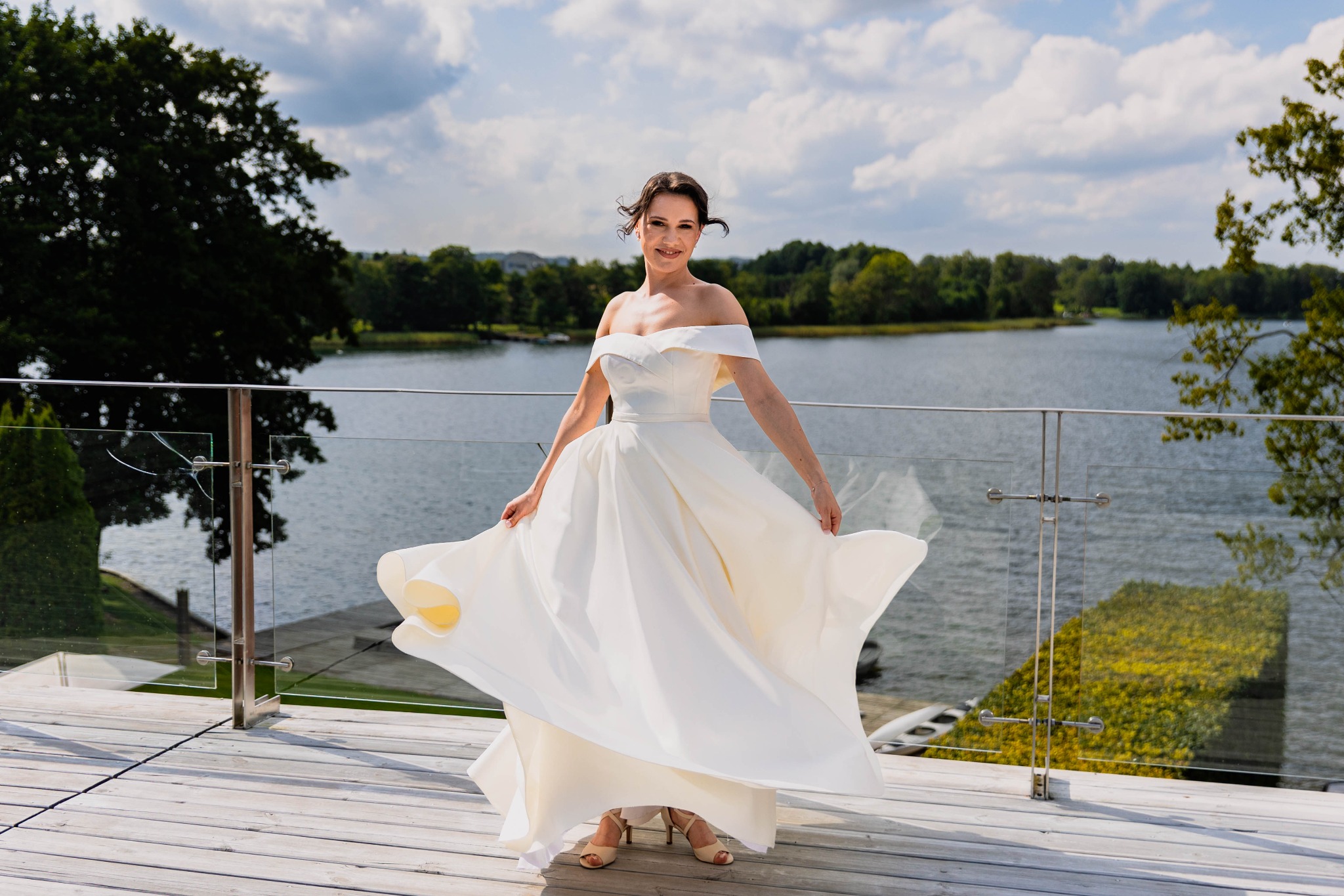 Uśmiechnięta panna młoda w białej sukni, rozłożonej w tańcu na drewnianym tarasie z widokiem na jezioro w słoneczny dzień. Romantyczna sceneria.