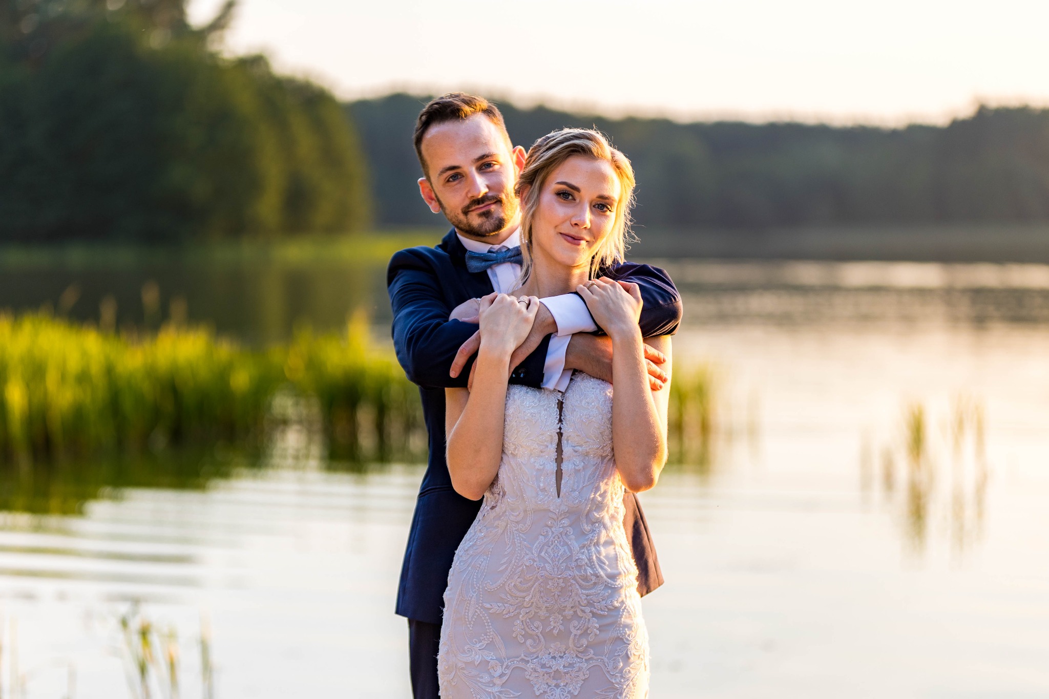 Elegancka para młoda w objęciach na tle jeziora o zachodzie słońca. Pan w granatowym garniturze i muszce, Panna Młoda w białej sukni z koronkowymi zdobieniami.