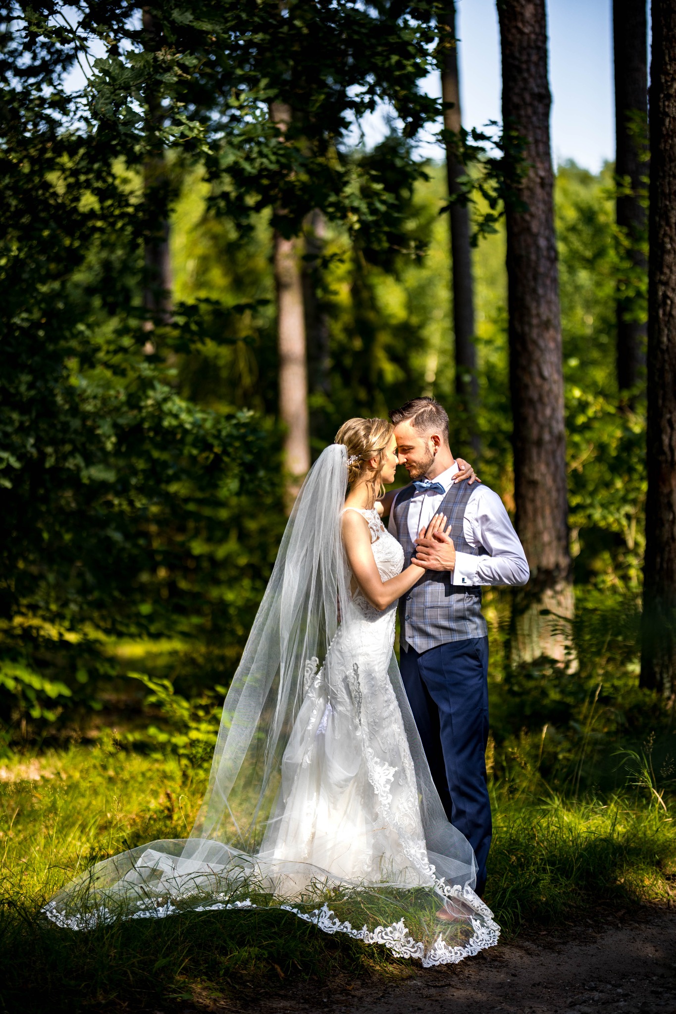 Para młoda w leśnej scenerii: panna młoda w koronkowej sukni i welonie, pan młody w eleganckiej kamizelce i muszce, delikatny dotyk dłoni, naturalne światło.