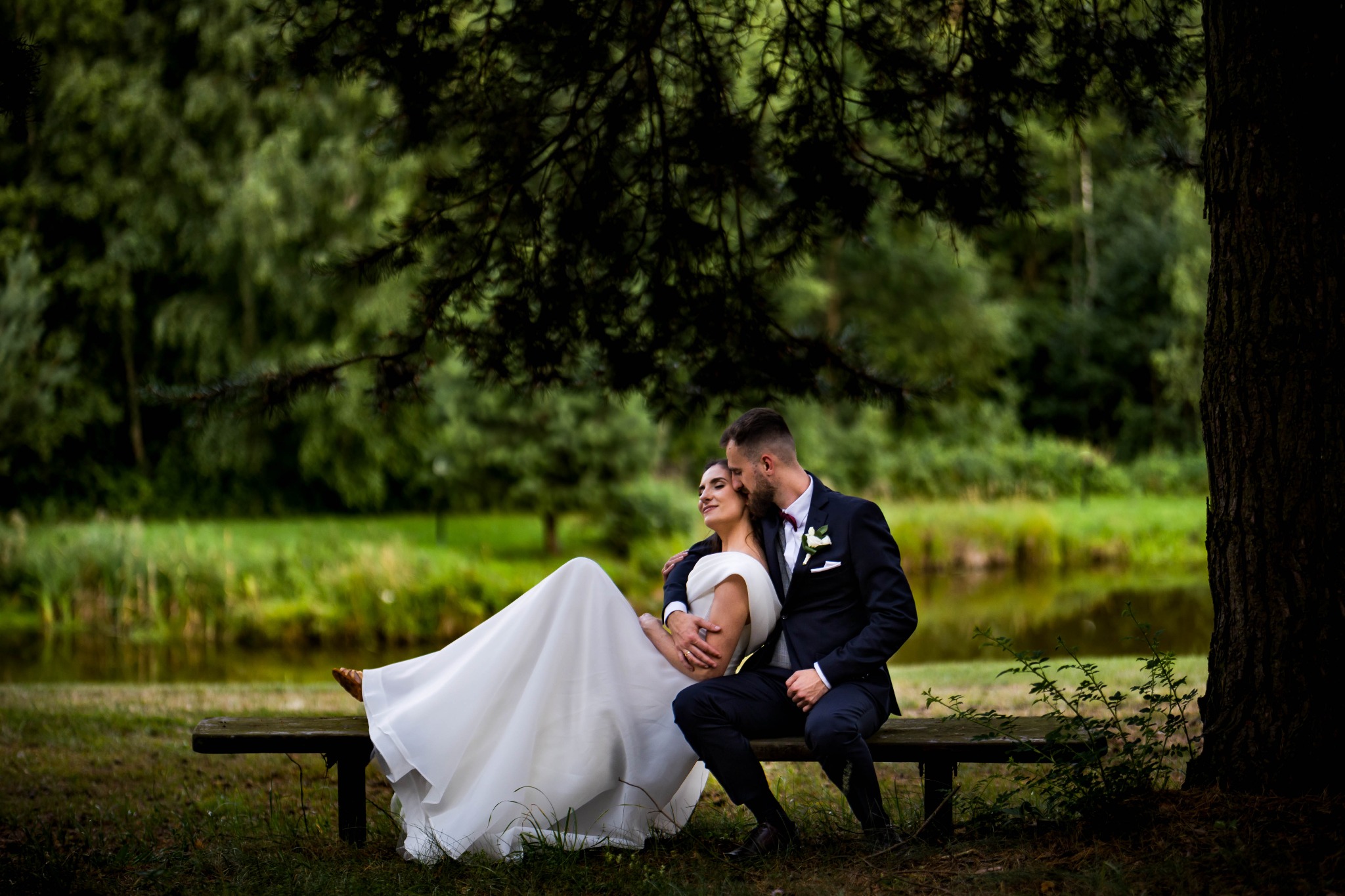 Para młoda w eleganckich strojach siedzi na ławce pod drzewem, w tle zielony krajobraz z jeziorem. Romantyczna sesja plenerowa w naturalnym otoczeniu.