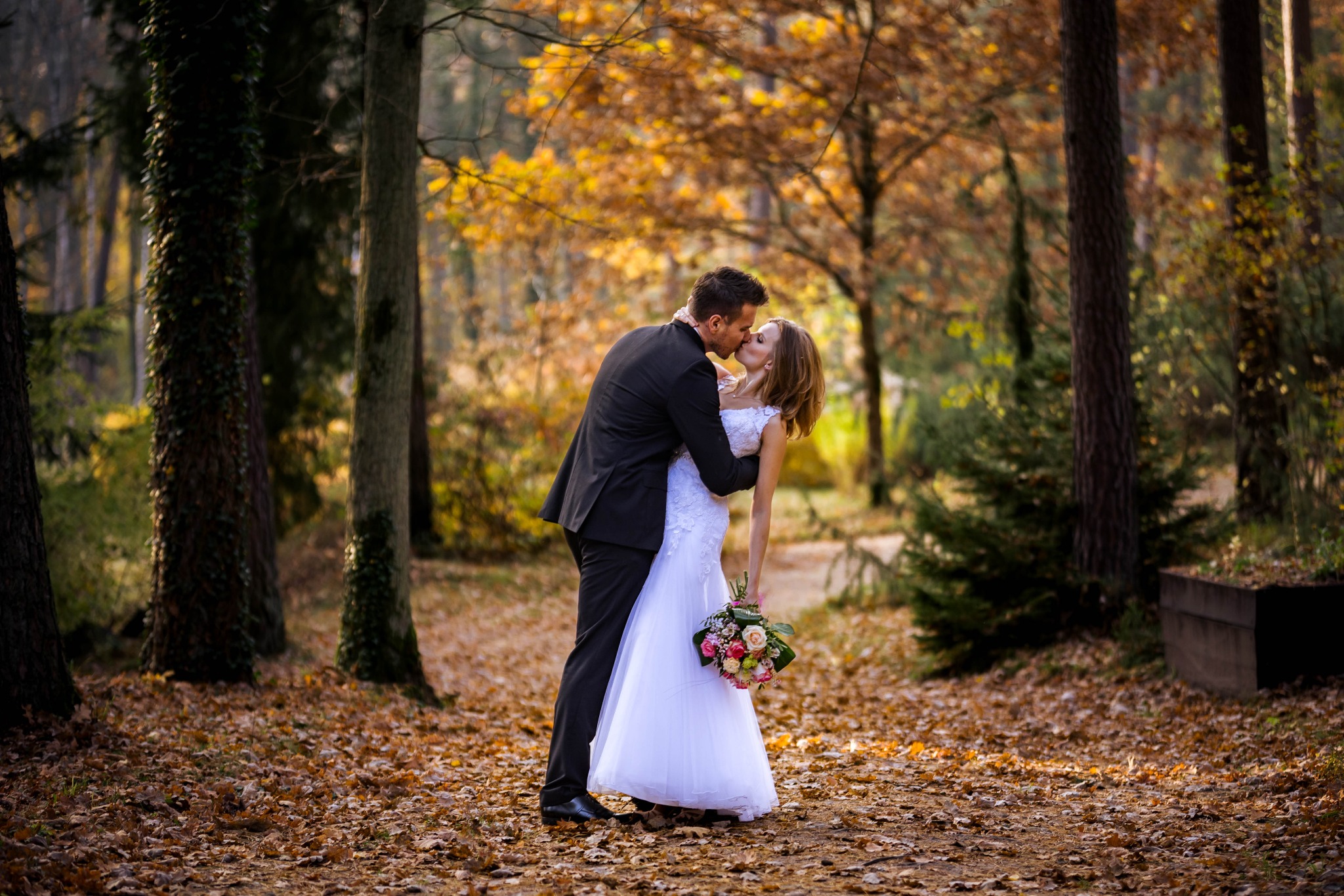 Jesienna sesja ślubna w lesie: Panna młoda w białej sukni i pan młody w czarnym garniturze całują się na tle drzew z żółtymi liśćmi, trzymając bukiet kwiatów.