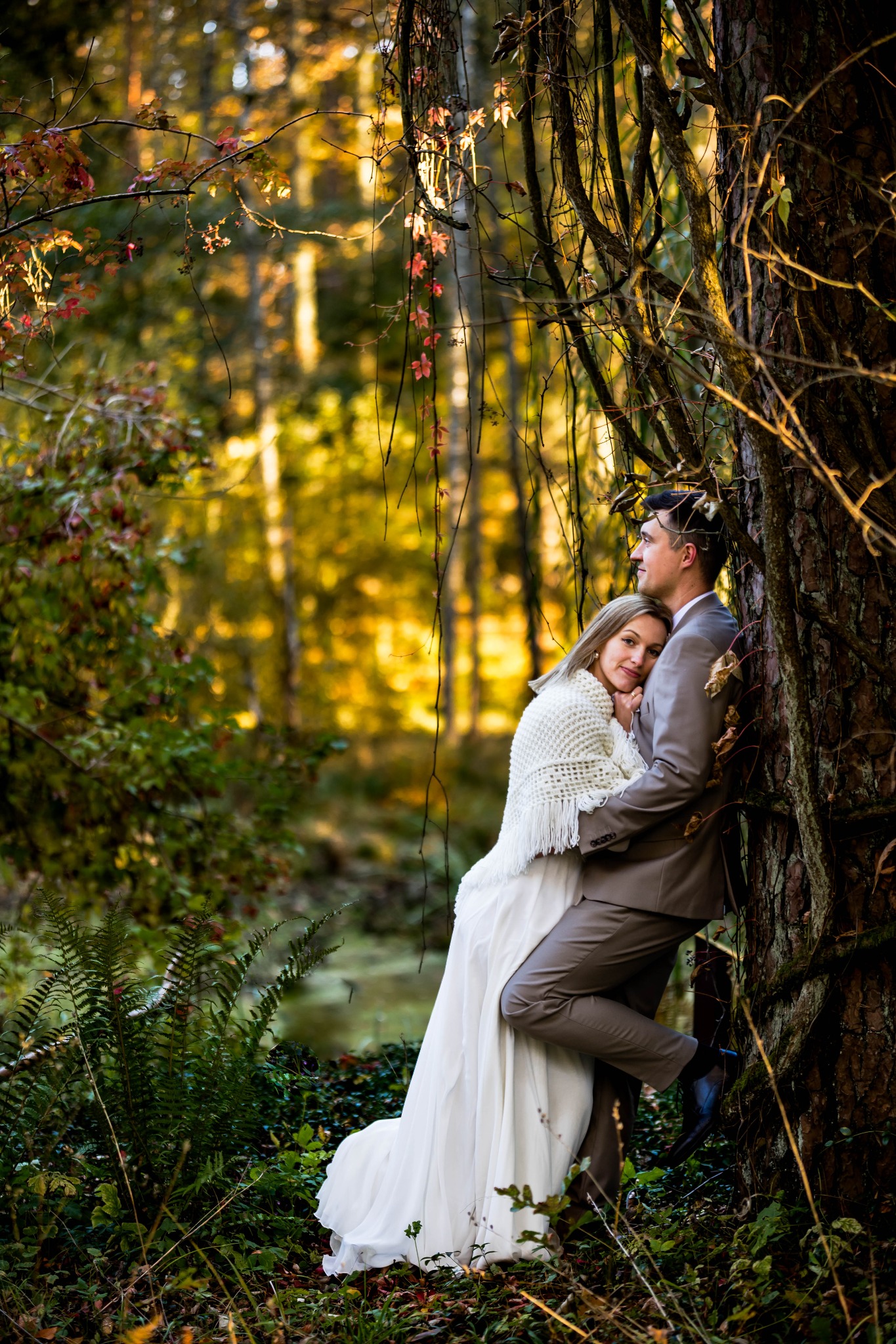 Para młoda w lesie, oparta o drzewo. Panna młoda w białej sukni i kremowym szalu, pan młody w szarym garniturze. Romantyczna sesja w naturalnym otoczeniu.