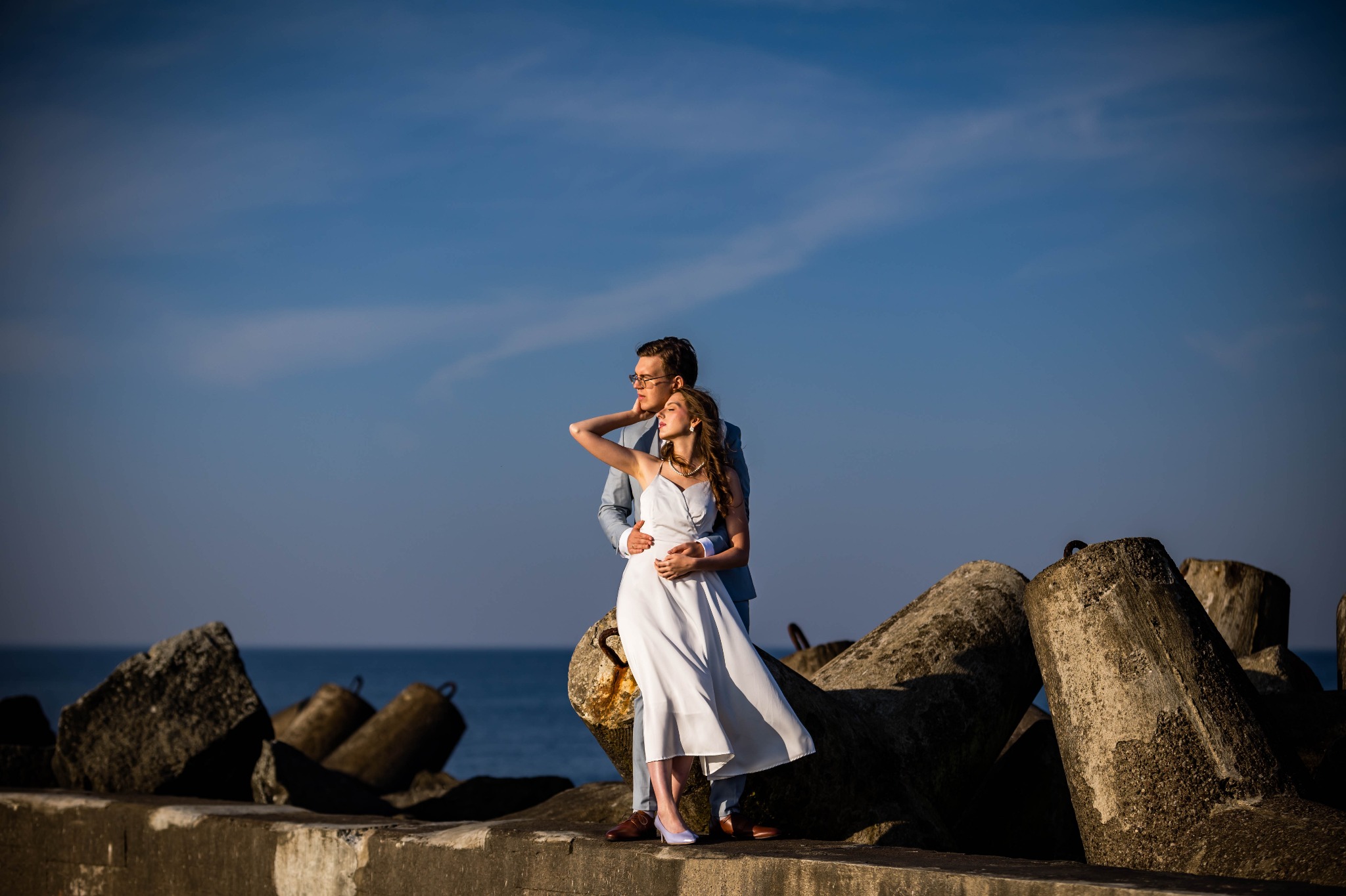 Para zakochanych na tle morza i nieba. Kobieta w białej sukience, mężczyzna w jasnym garniturze. Ujęcie na betonowym nabrzeżu z falochronami.