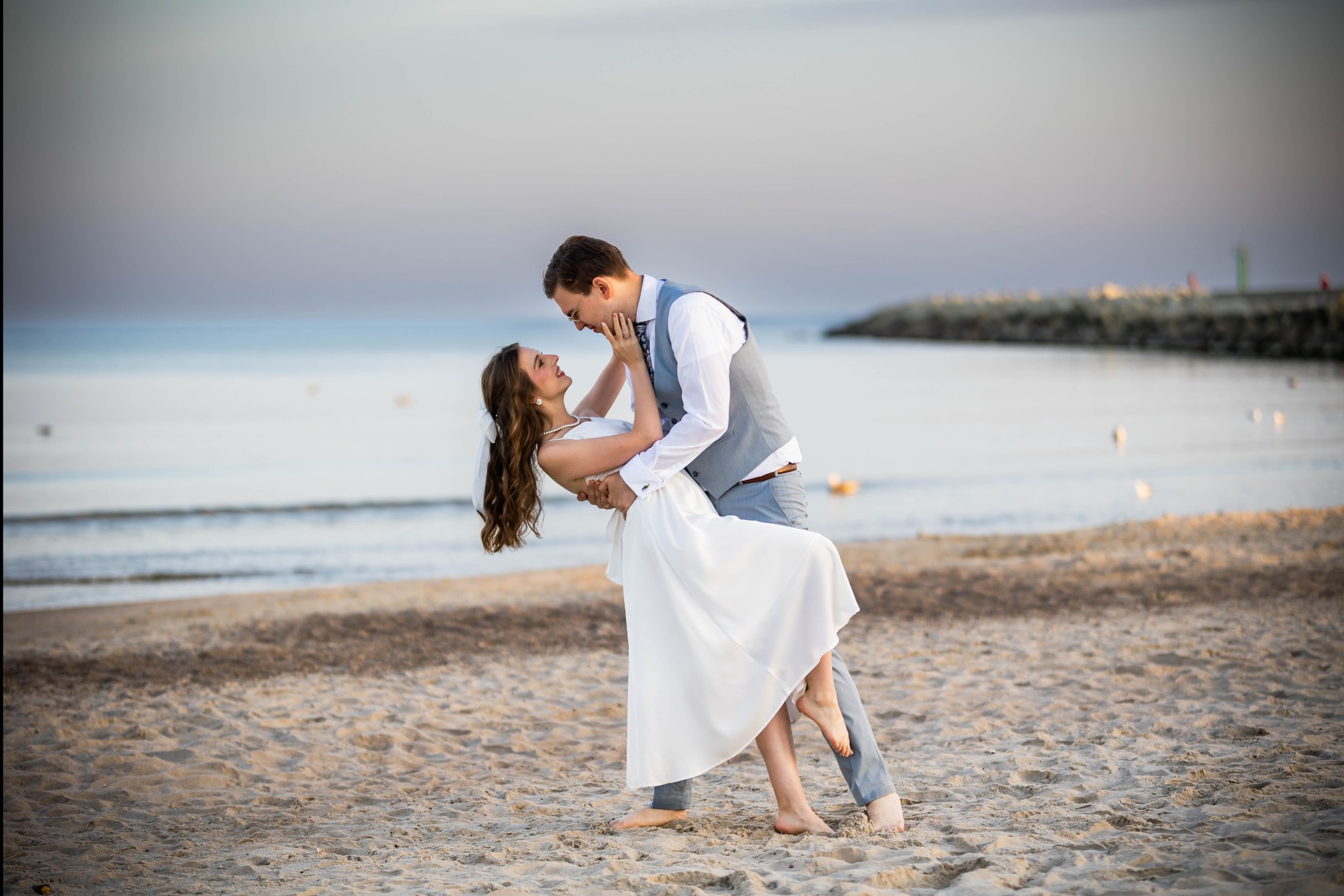 Para młoda tańczy boso na piaszczystej plaży o zmierzchu. Ona w białej sukni, on w szarej kamizelce. W tle woda i falochron. Romantyczna sesja ślubna.