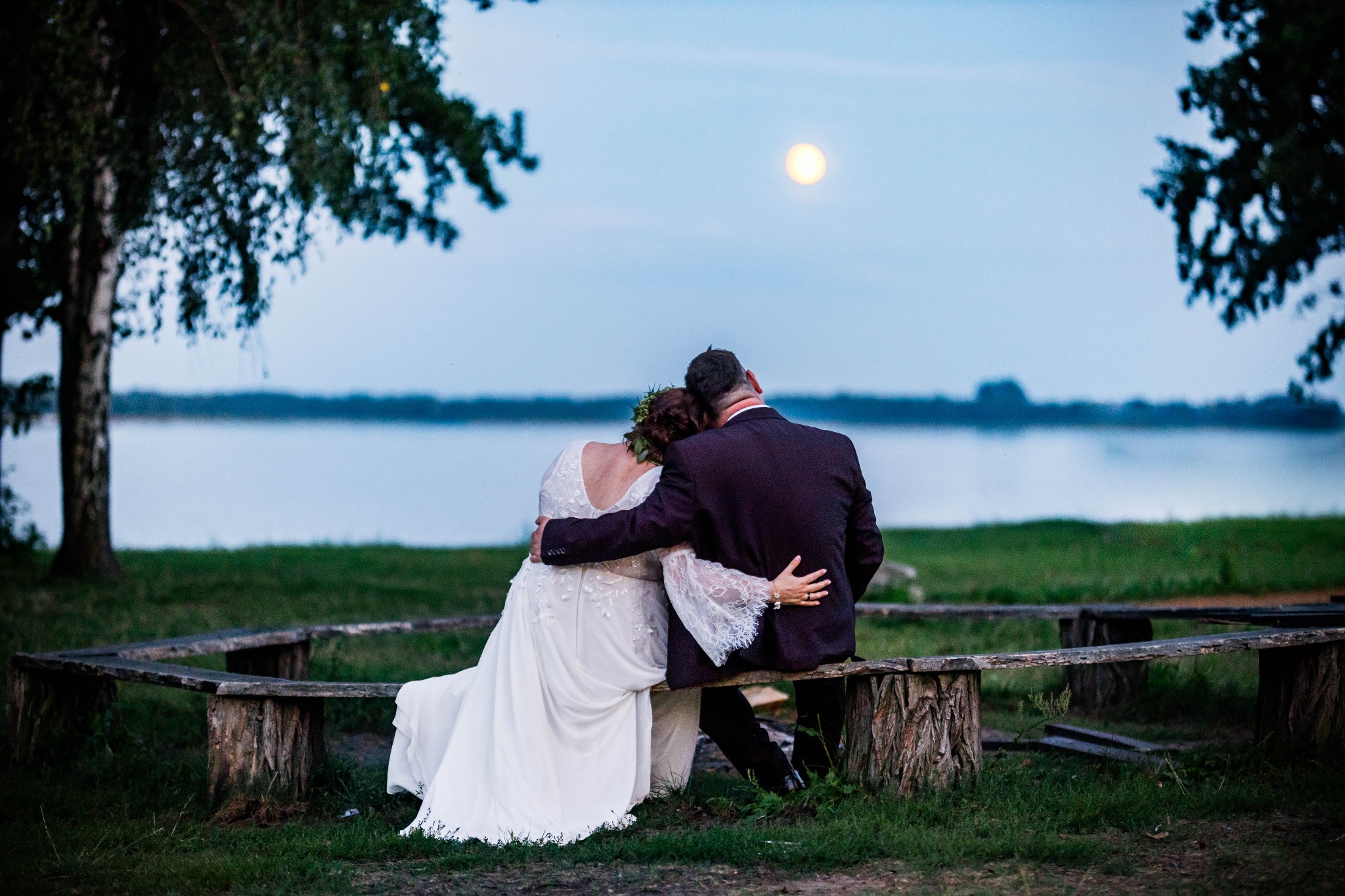 Para młoda siedzi na ławce z pni drzew, obejmując się na tle jeziora i księżyca w pełni. Suknia panny młodej z koronkowymi rękawami. Ujęcie z tyłu, wieczorny plener.