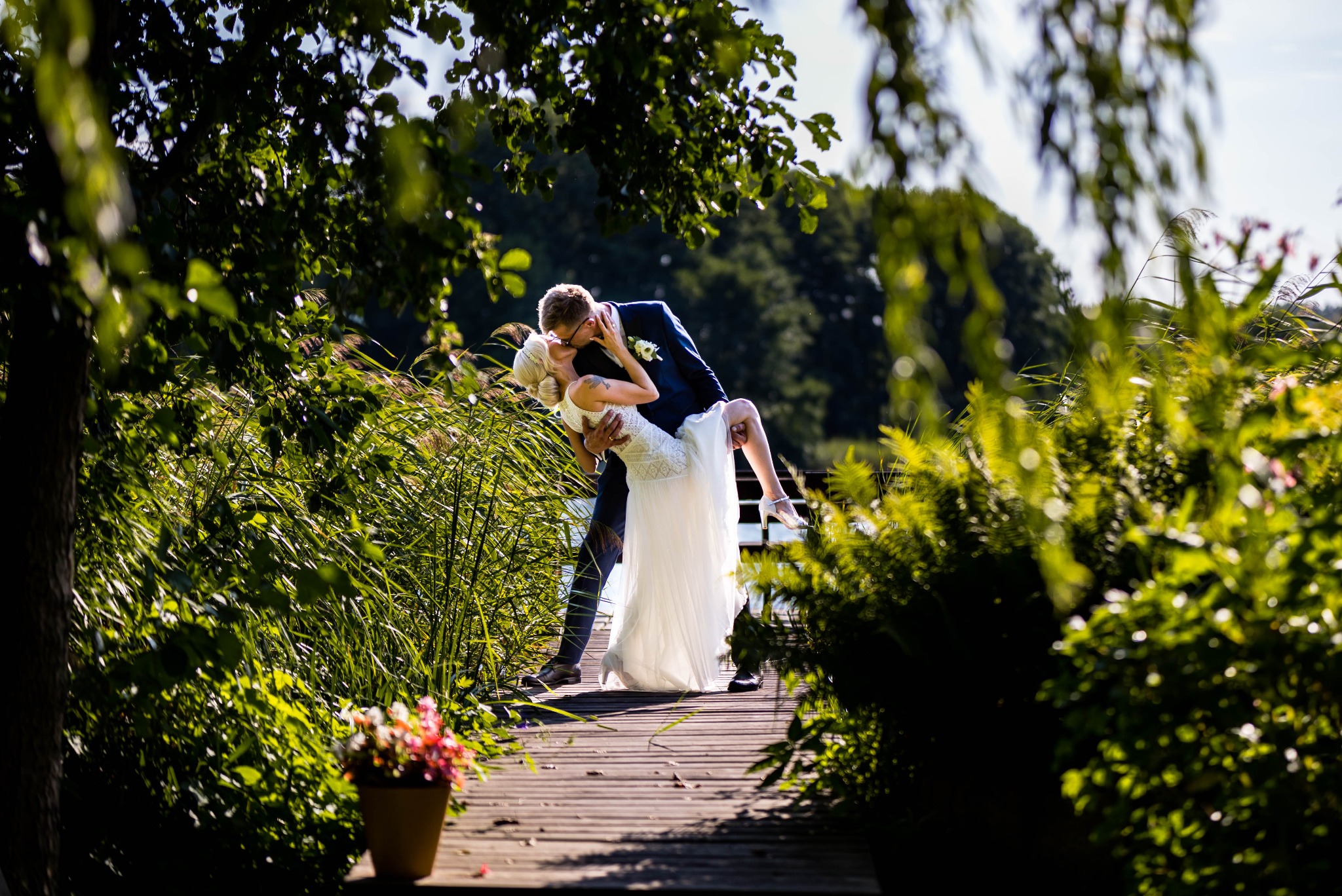 Para młoda w tańcu na drewnianym pomoście, otoczona zielenią. Pan młody w granatowym garniturze, panna młoda w białej sukni. Romantyczna sceneria, naturalne światło.