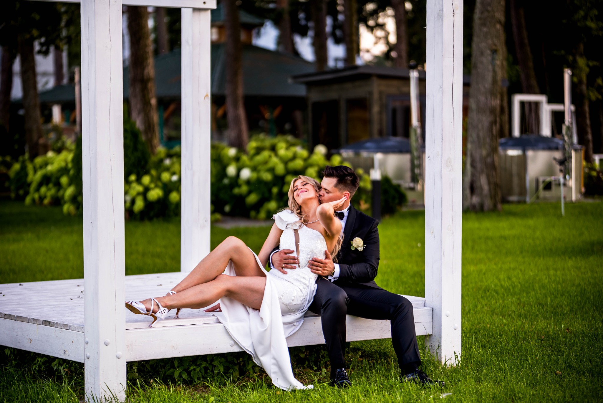 Para młoda w objęciach na białej, drewnianej platformie w plenerze. Panna młoda w białej sukni, pan młody w czarnym smokingu. Romantyczna atmosfera, naturalne światło.