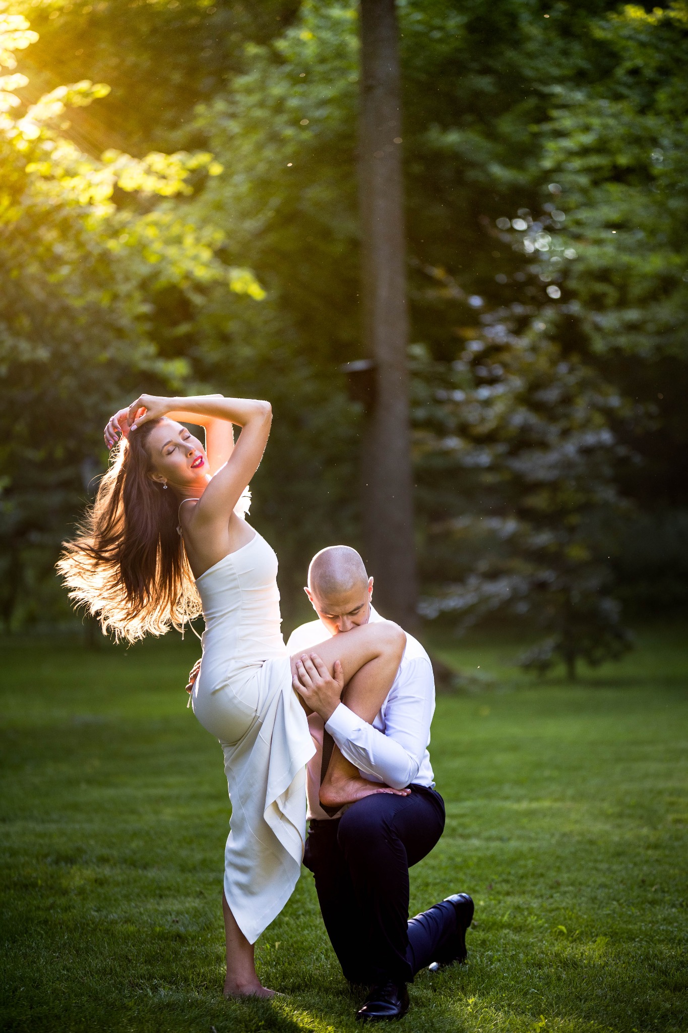 Romantyczna sesja w plenerze: mężczyzna klęczy, całując nogę kobiety w białej sukni na tle zieleni i promieni słonecznych. Dynamiczne ujęcie, podkreślone światłem.