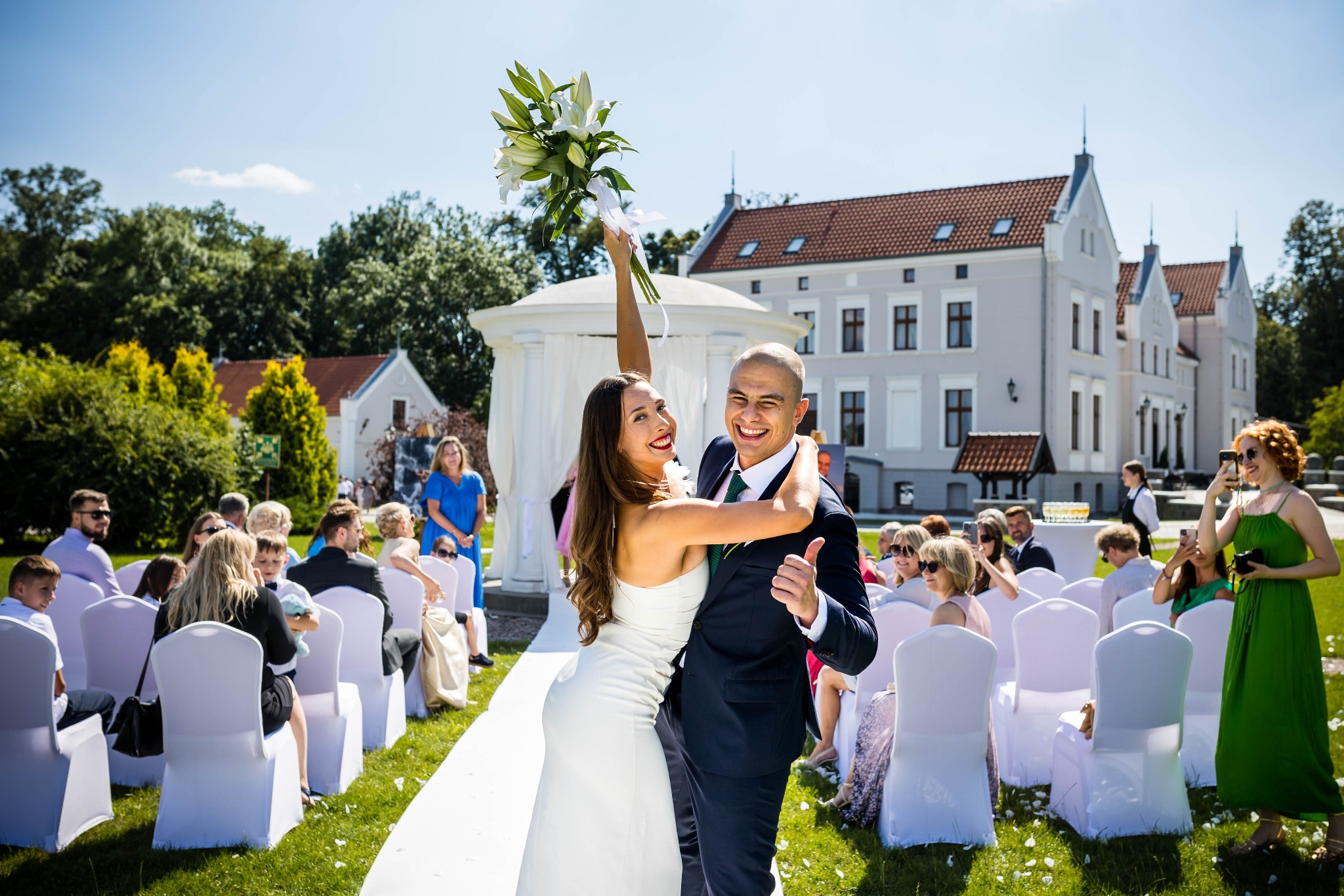 Uśmiechnięta para młoda w objęciach na tle białej ścieżki, gości i zabytkowego budynku w słoneczny dzień. Panna młoda trzyma bukiet lilii.