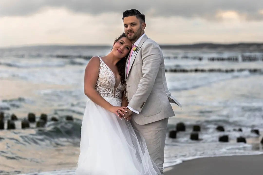 Para młoda na plaży: Panna młoda w koronkowej sukni, Pan młody w beżowym garniturze. Romantyczny kadr z sesji ślubnej na tle morza i drewnianych pali.