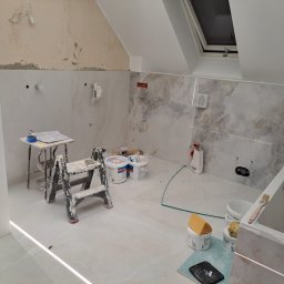 usługi budowlano-wykończeniowe - Łazienka w trakcie wykańczania: częściowo położone marmurowe płytki na ścianach, widoczne narzędzia glazurnicze, drabina malarska i puste wiadra po kleju na podłodze, sufit skośny z oknem dachowym.