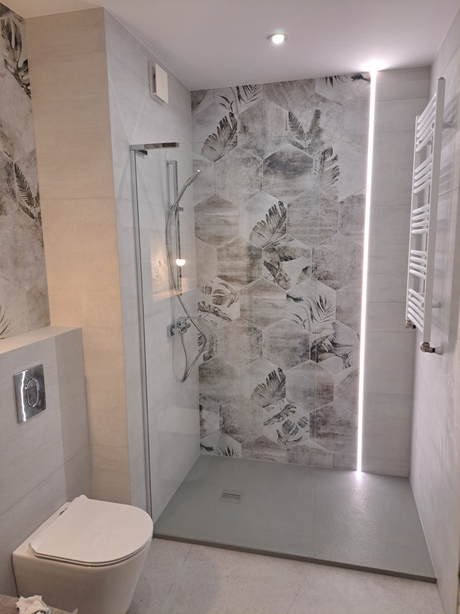Nowoczesna łazienka z kabiną typu walk-in, heksagonalnymi płytkami z motywem liści na ścianie prysznicowej, podświetleniem LED w pionowej szczelinie i wiszącą toaletą.