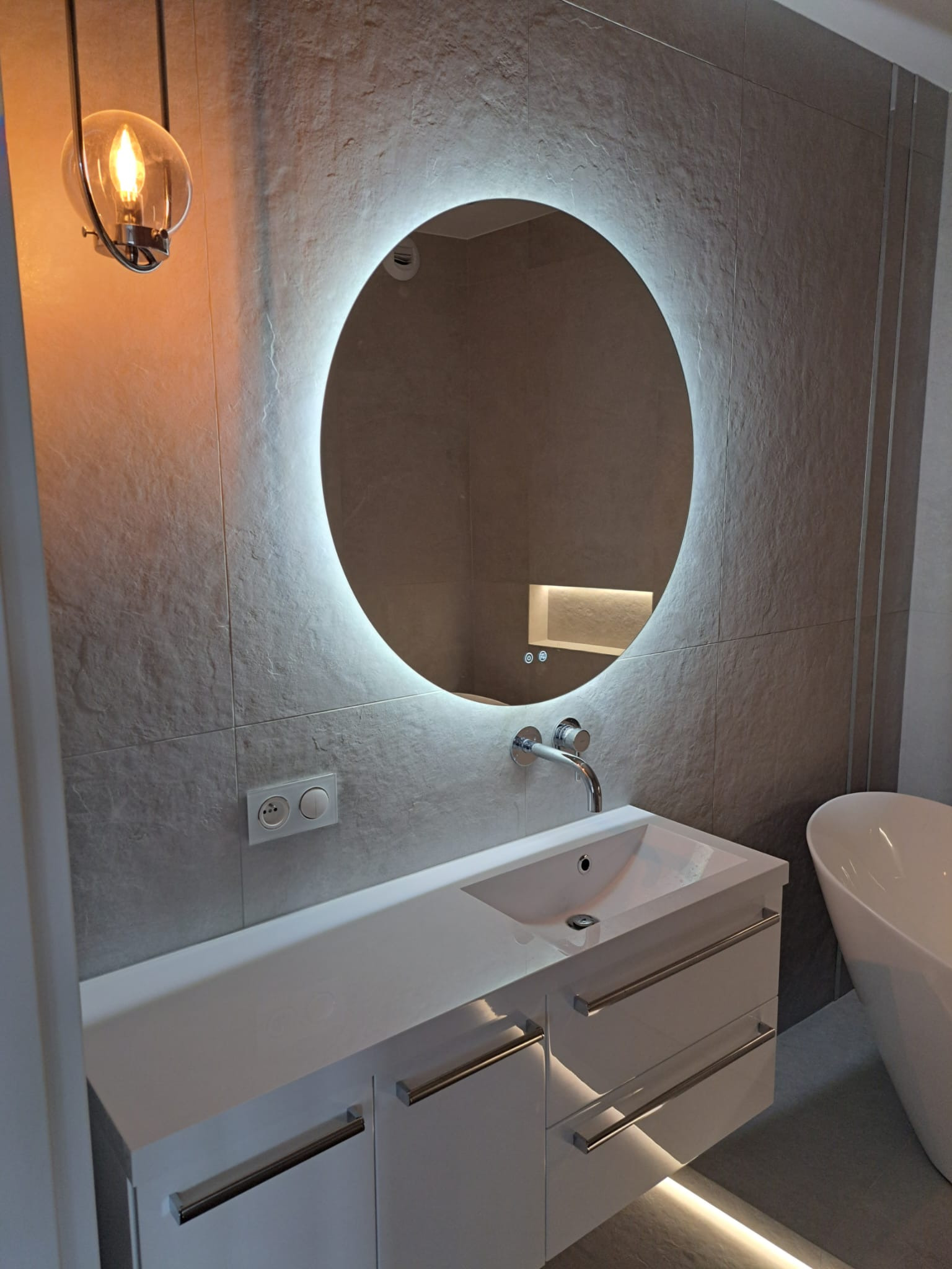 Nowoczesna łazienka z podświetlanym okrągłym lustrem, białą szafką pod umywalką z chromowanymi uchwytami, szarą kamienną okładziną ścienną i wolnostojącą wanną.