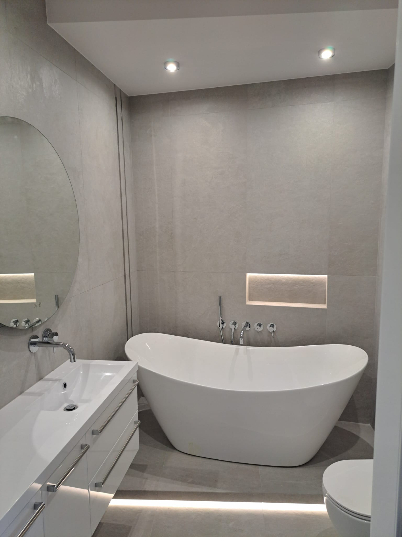 Nowoczesna łazienka w odcieniach szarości z wolnostojącą białą wanną, okrągłym lustrem, podświetlaną wnęką i podłogowym oświetleniem LED.