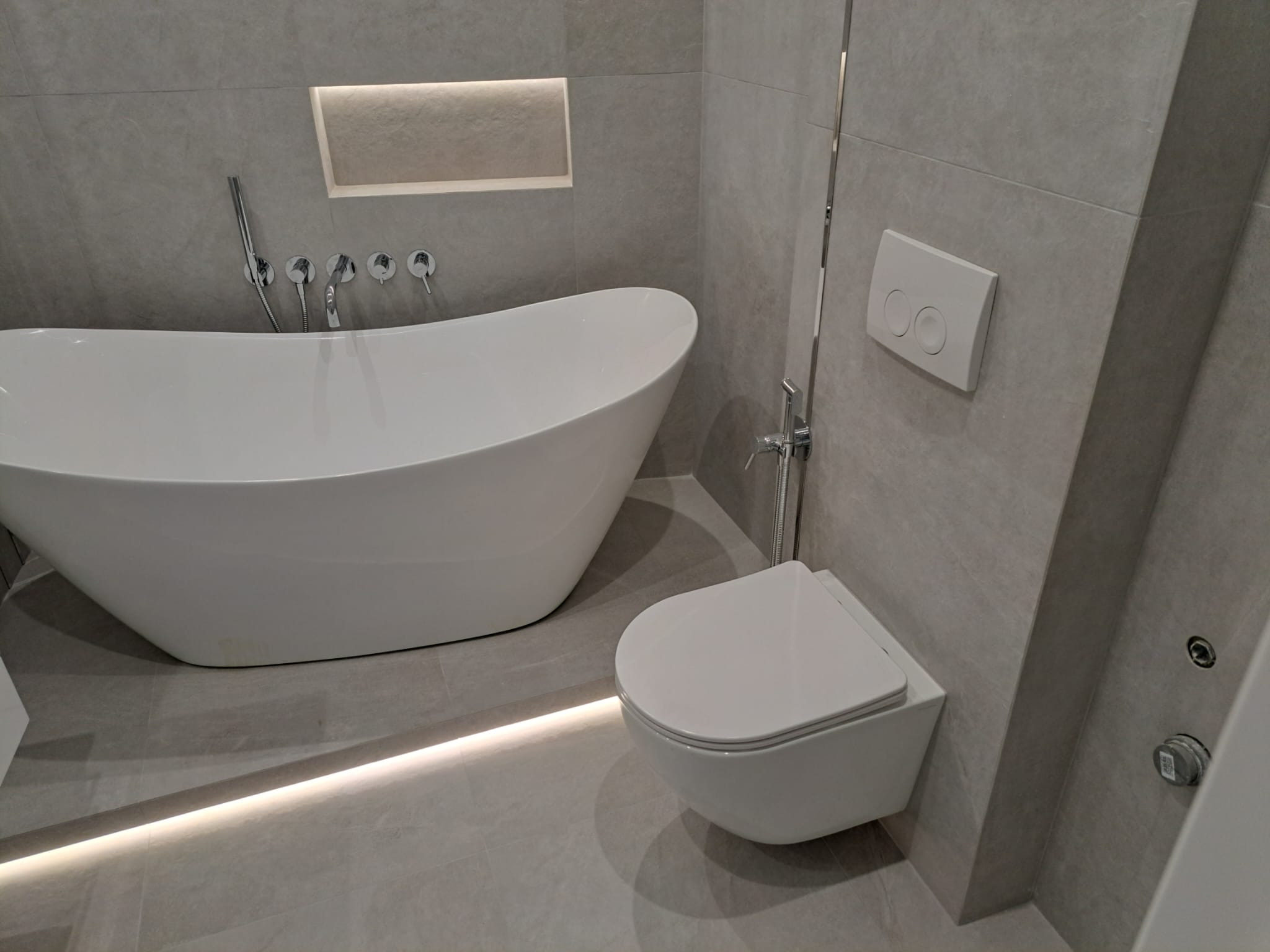 Nowoczesna łazienka w odcieniach szarości z wolnostojącą białą wanną, podwieszanym WC i subtelnym oświetleniem LED w podłodze oraz wnęce ściennej nad wanną.