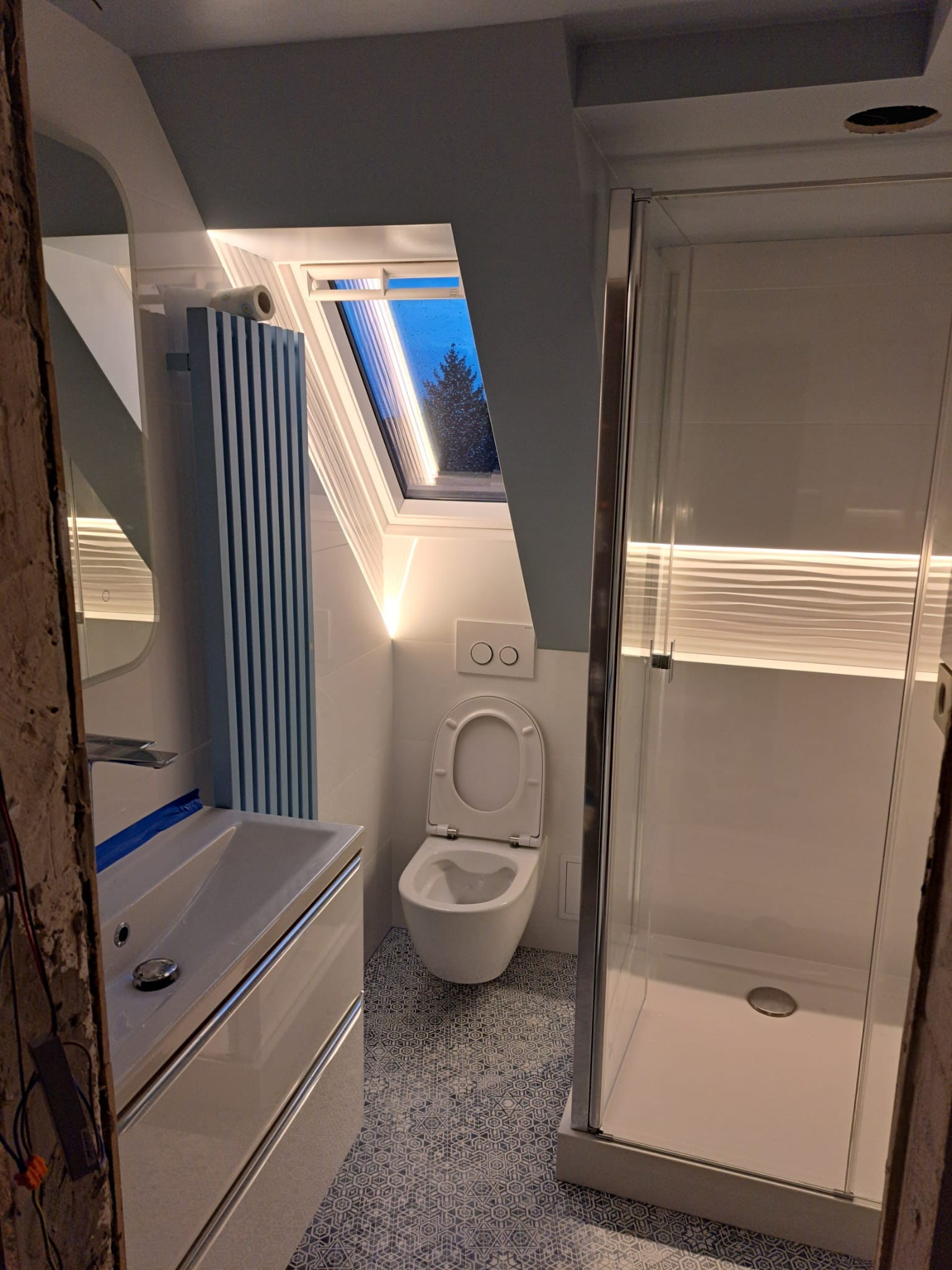 Wykończona łazienka na poddaszu z kabiną prysznicową, toaletą, umywalką z szafką i grzejnikiem żeberkowym w niebieskie paski, oświetlona paskami LED pod sufitem i nad kabiną prysznicową, z oknem...