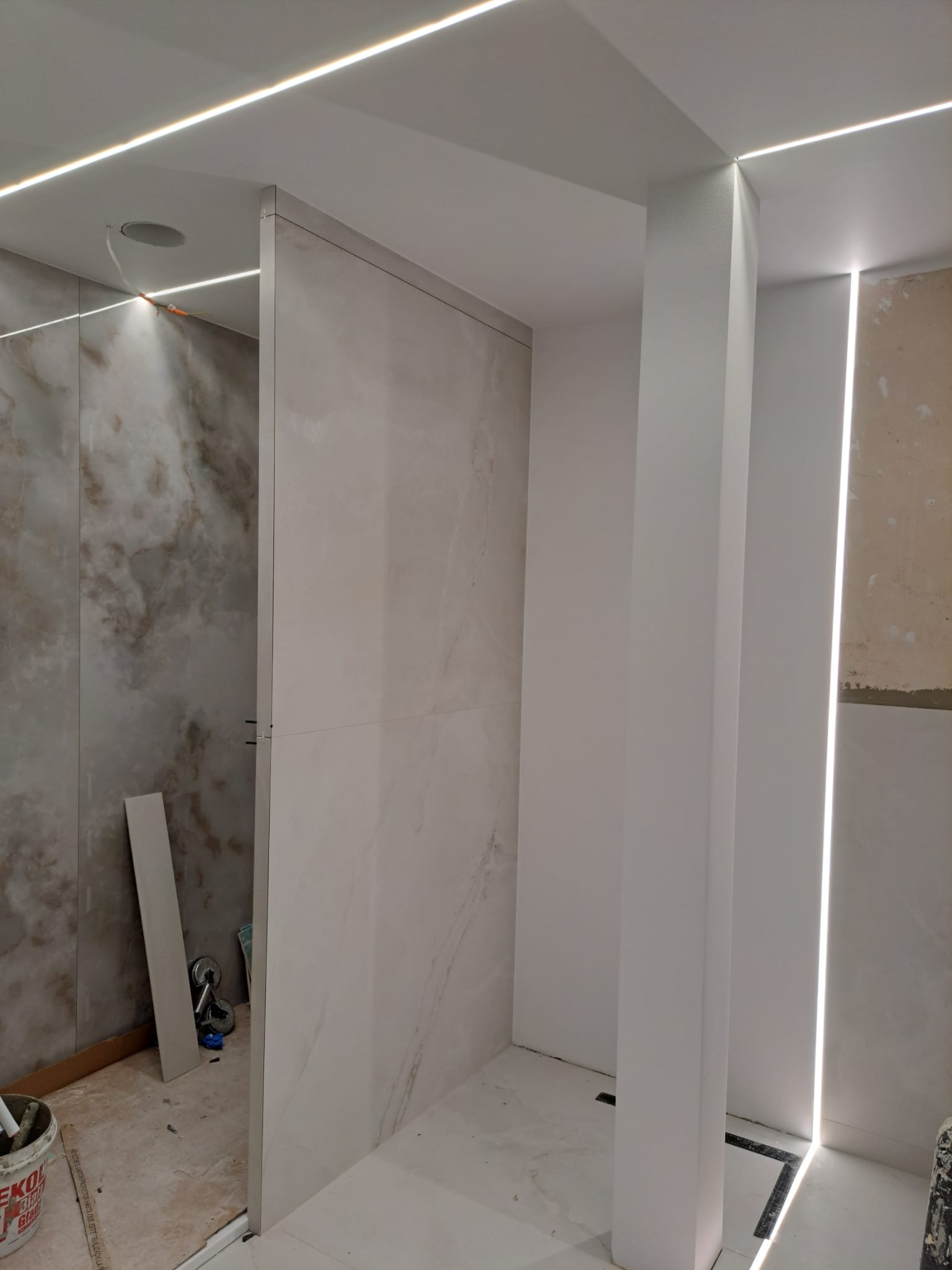 Wnętrze łazienki w trakcie wykańczania z marmurowymi płytkami na ścianach, oświetleniem LED w suficie i kolumną obudowaną płytami gipsowo-kartonowymi.