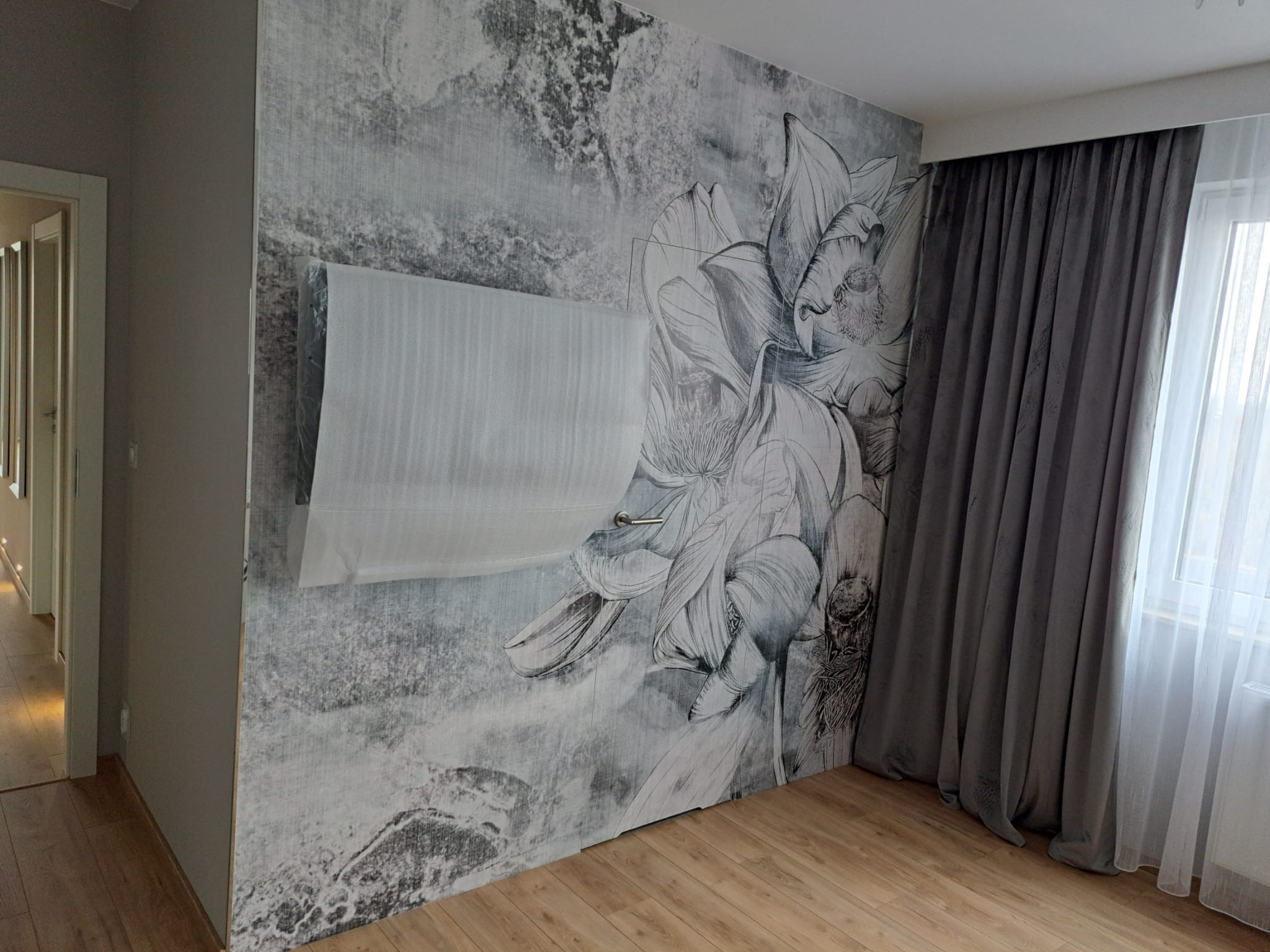 Pokój z fototapetą w kwiaty, zasłoniętym oknem roletą w folii ochronnej i drewnianą podłogą.