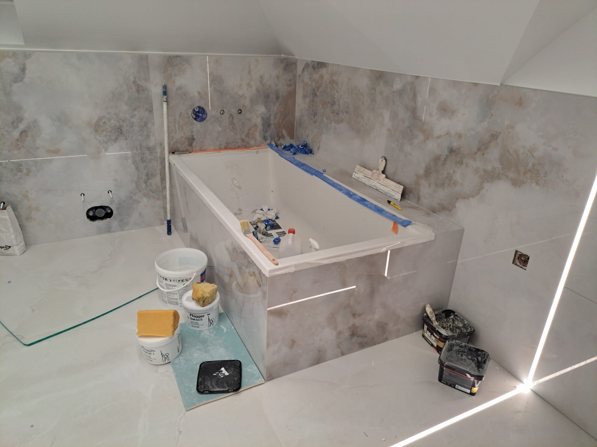 Wnętrze łazienki w trakcie układania płytek, widoczna wanna obudowana płytkami imitującymi marmur, narzędzia glazurnicze, pędzle i pojemniki z klejem.