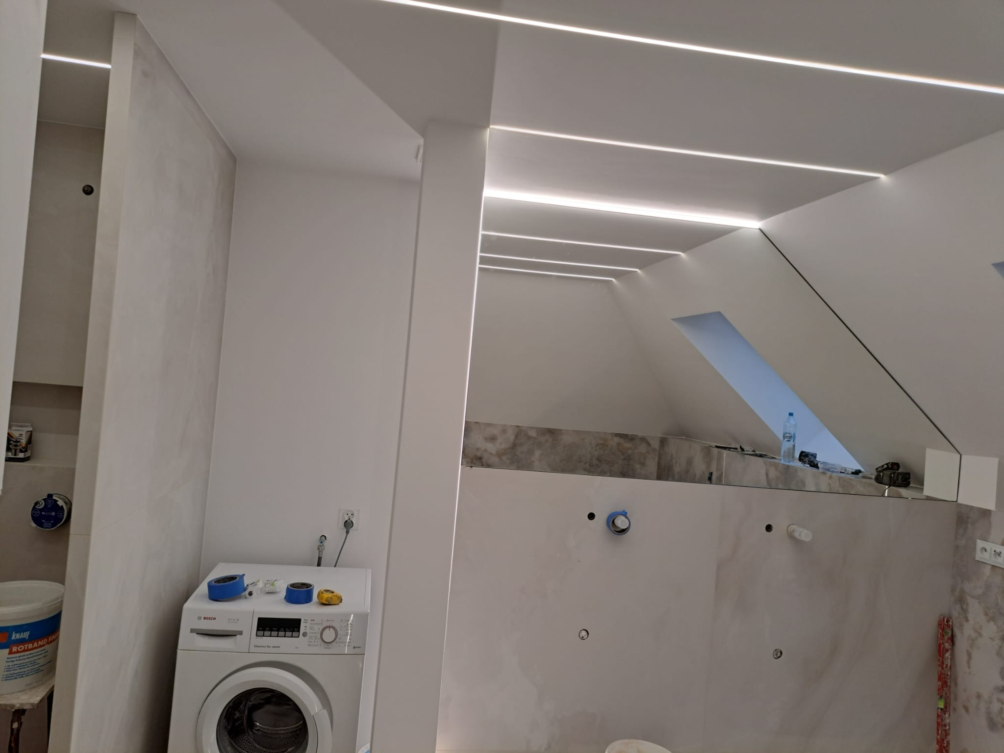Remont łazienki na poddaszu: Białe ściany, płytki imitujące marmur, pralka Bosch z taśmą malarską na górze, oświetlenie sufitowe LED w profilach.