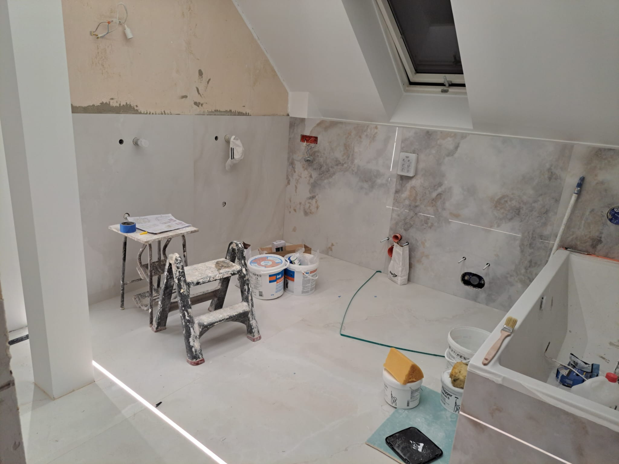 Łazienka w trakcie wykańczania: częściowo położone marmurowe płytki na ścianach, widoczne narzędzia glazurnicze, drabina malarska i puste wiadra po kleju na podłodze, sufit skośny z oknem dachowym.