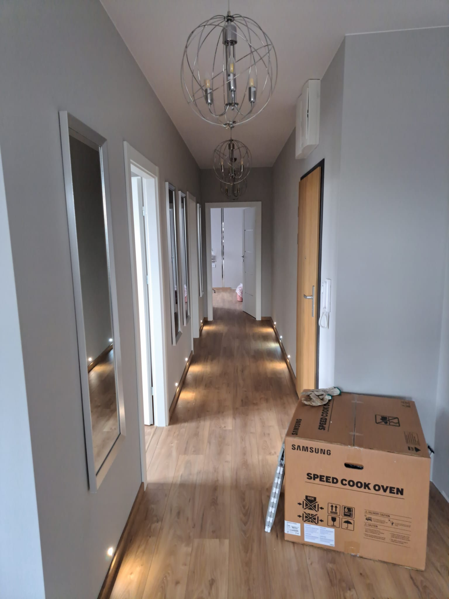 Jasny korytarz z panelami podłogowymi, oświetleniem punktowym wzdłuż ścian, lustrami i wiszącymi lampami kulistymi, z kartonem po piekarniku Samsung Speed Cook Oven na podłodze.
