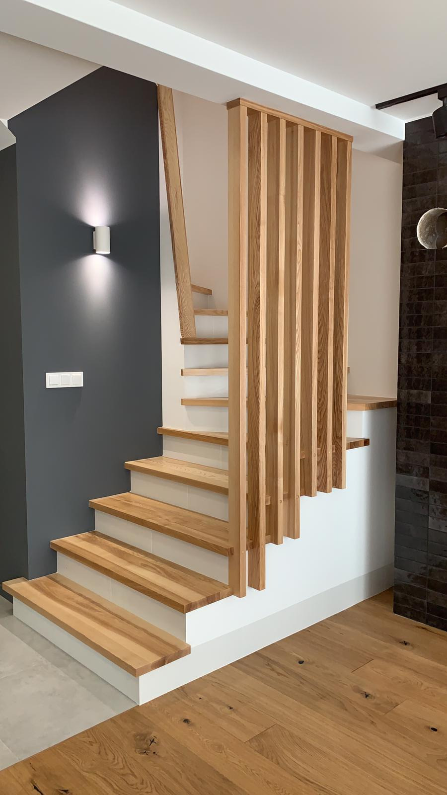 Nowoczesne schody drewniane z ażurową balustradą, oświetlone kinkietem na ciemnoszarej ścianie, z widoczną podłogą z desek.