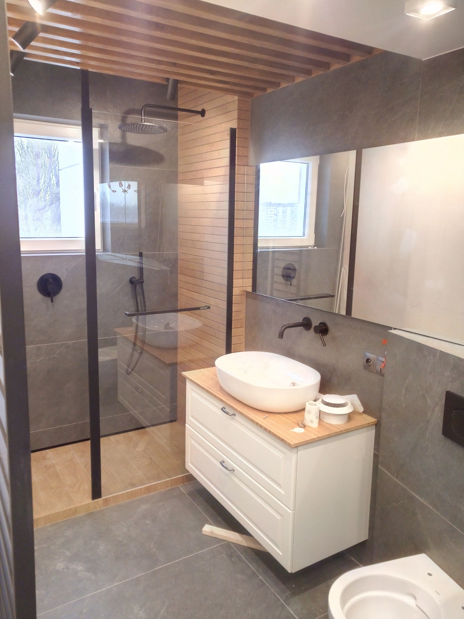Nowoczesna łazienka z kabiną prysznicową typu walk-in, umywalką nablatową na szafce z drewnianym blatem, szarą glazurą i drewnianym sufitem.