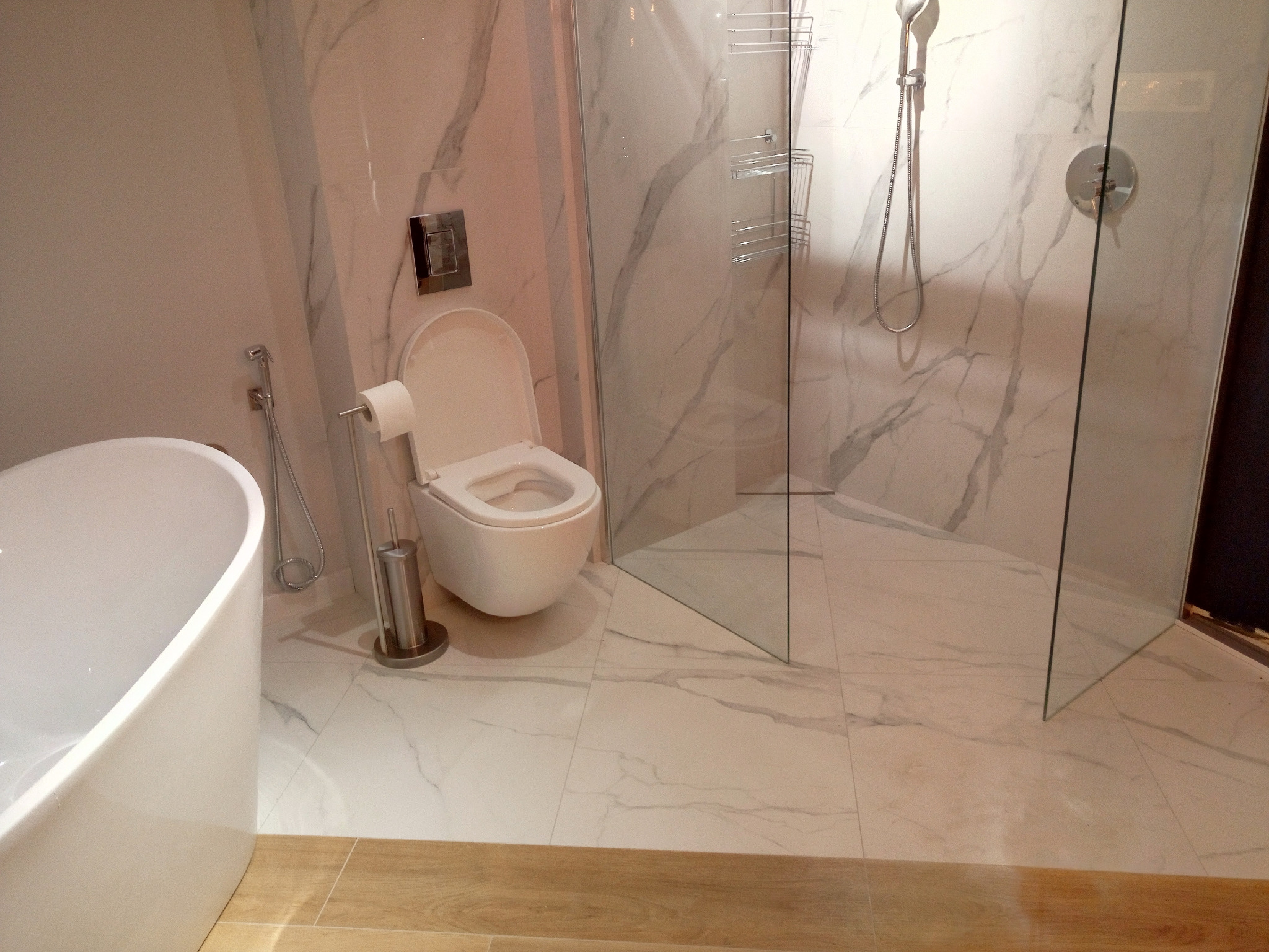 Nowoczesna łazienka z marmurowymi płytkami, widoczna wanna wolnostojąca, toaleta z bidetką i kabina prysznicowa ze szklanymi ściankami.