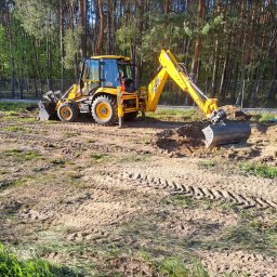 Rybołowik Prace Ziemne - Żółta koparko-ładowarka JCB kopie dół w ziemi na tle lasu i ogrodzenia z siatki, widoczne świeże ślady opon na piasku.