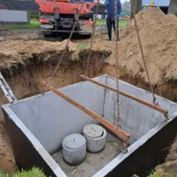 Betonowy zbiornik z dwoma pokrywami opuszczany do wykopu przy użyciu dźwigu samochodowego, pracownik w oddali nadzoruje proces.