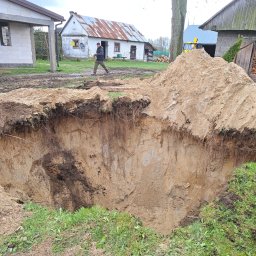 Rybołowik Prace Ziemne - Głęboki wykop w piaszczystej glebie na wiejskim terenie, z pryzmą ziemi obok, w tle budynki gospodarcze i przechodzący mężczyzna z pilarką.