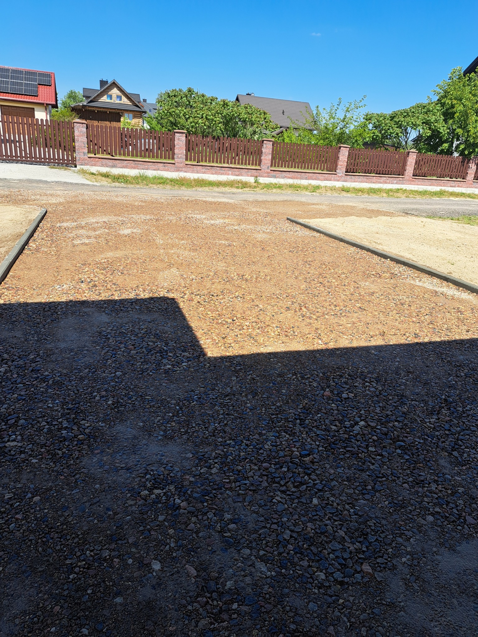 Przygotowany teren pod budowę, z widocznymi obrzeżami i częściowo utwardzoną nawierzchnią z kruszywa, w tle ogrodzenie posesji i zabudowa jednorodzinna w słoneczny dzień.