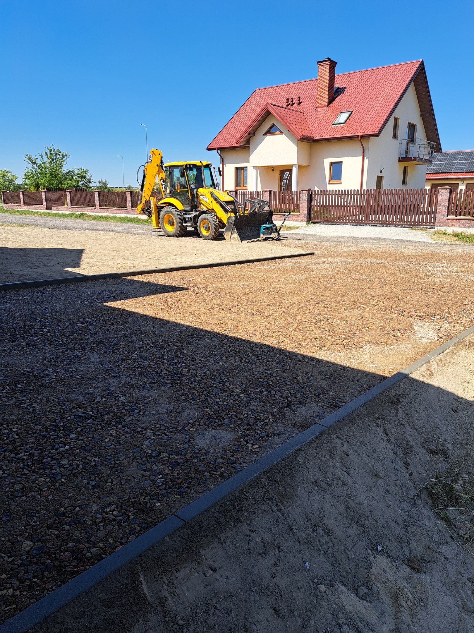 Żółta koparko-ładowarka na placu budowy przed domem jednorodzinnym, teren przygotowany pod kostkę brukową, widoczne obrzeża i podsypka.