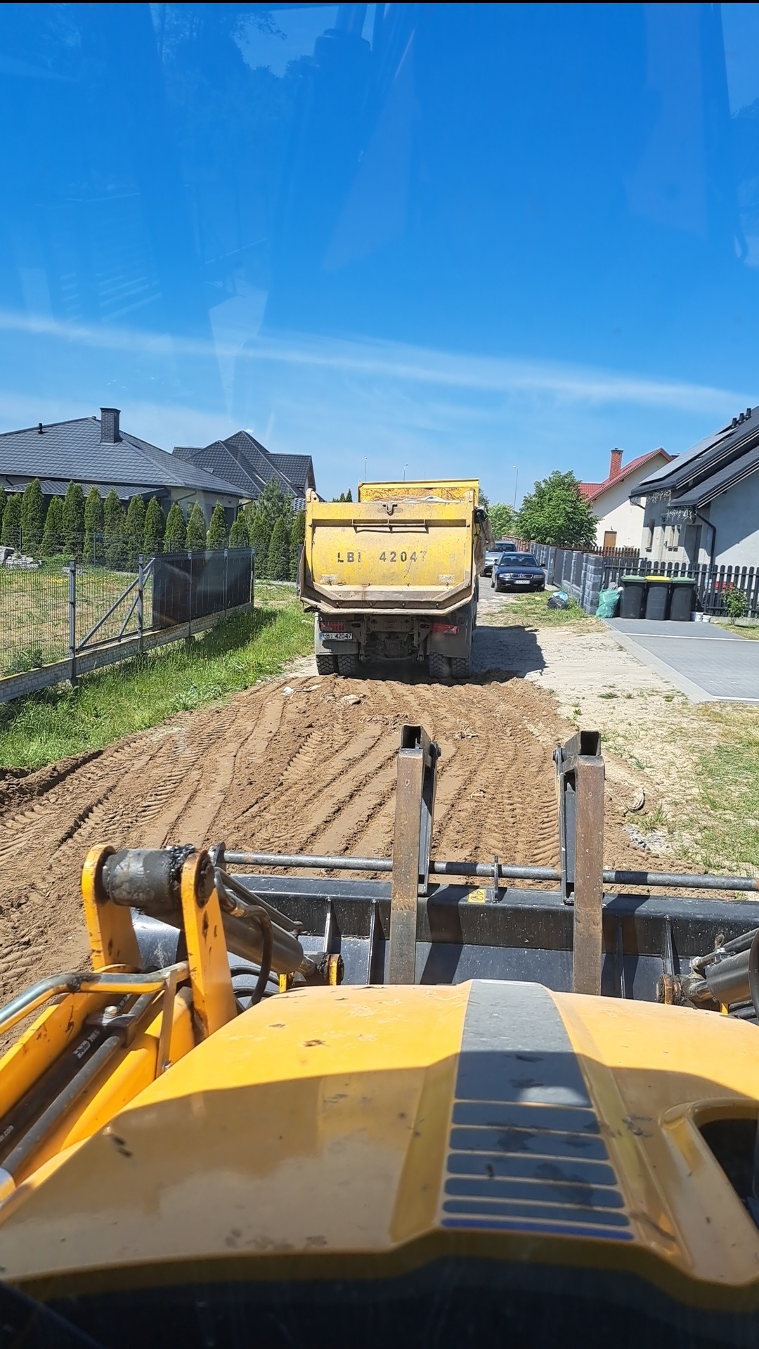 Widok z kabiny ładowarki na żółtą wywrotkę z numerem LBI 42047 na drodze gruntowej, w tle domy jednorodzinne i błękitne niebo.
