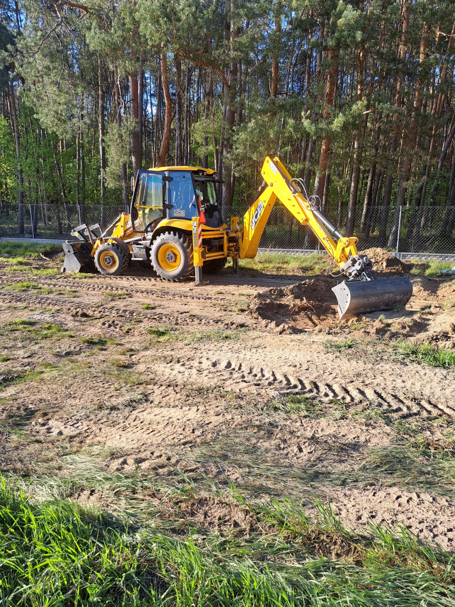 Żółta koparko-ładowarka JCB kopie dół w ziemi na tle lasu i ogrodzenia z siatki, widoczne świeże ślady opon na piasku.