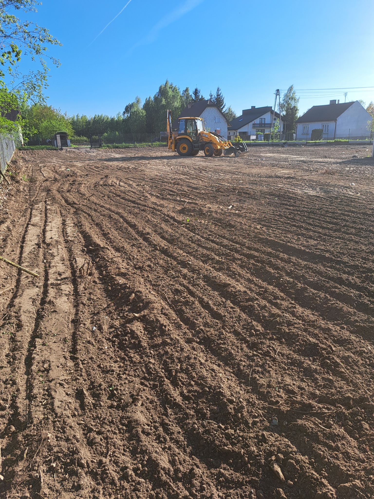 Żółta koparko-ładowarka na wyrównanym, brązowym terenie budowlanym, widoczne ślady opon, w tle domy jednorodzinne i drzewa pod błękitnym niebem.