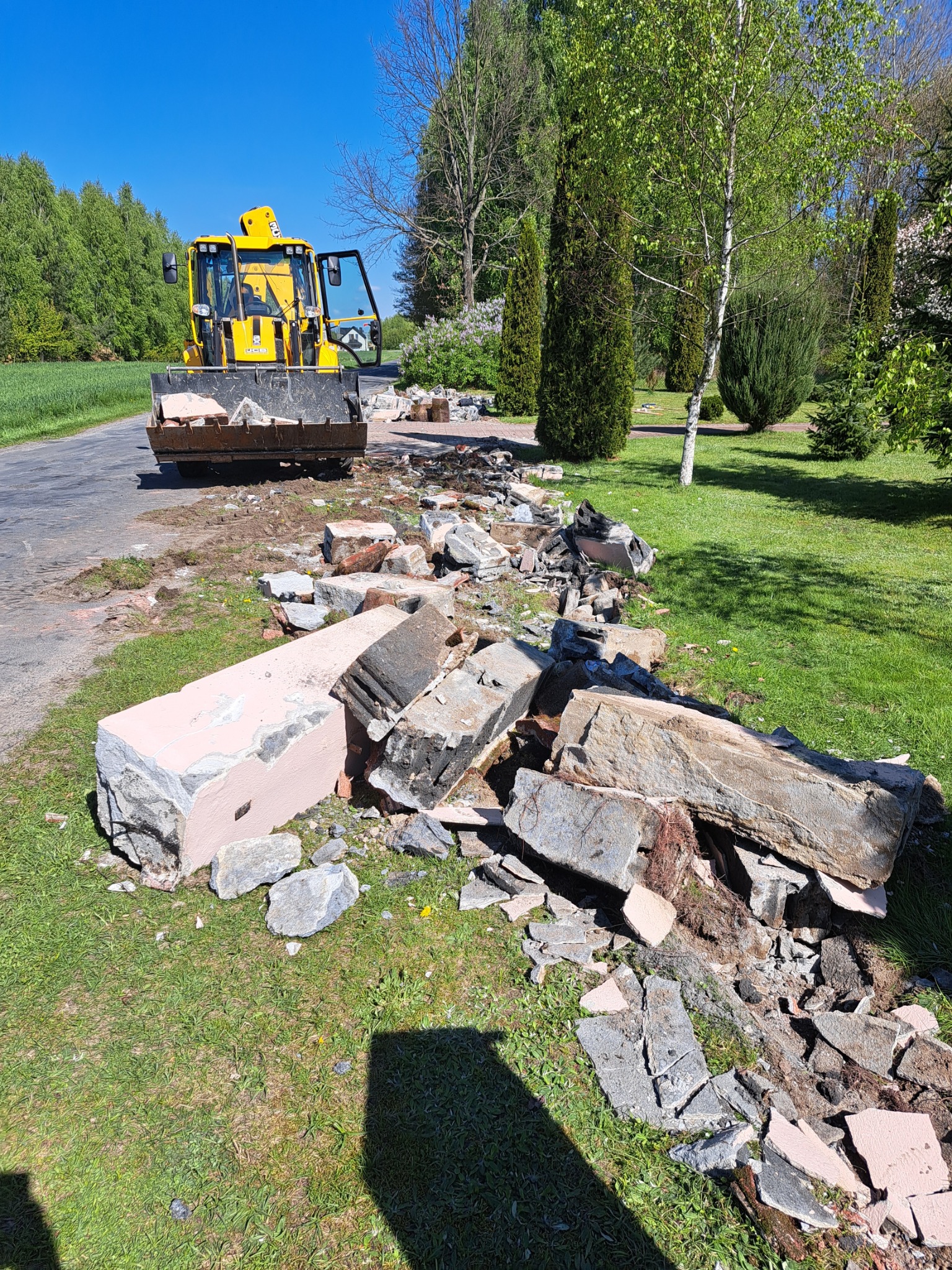 Żółta ładowarka czołowa z otwartymi drzwiami stoi na asfalcie obok sterty gruzu i fragmentów rozbitego betonu na trawniku, w tle drzewa i błękitne niebo.