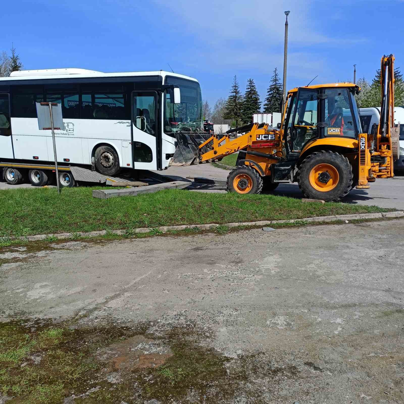 Żółta koparko-ładowarka JCB podnosi uszkodzony autobus na platformę transportową na placu, widoczne trawniki, drzewa iglaste i słup oświetleniowy.