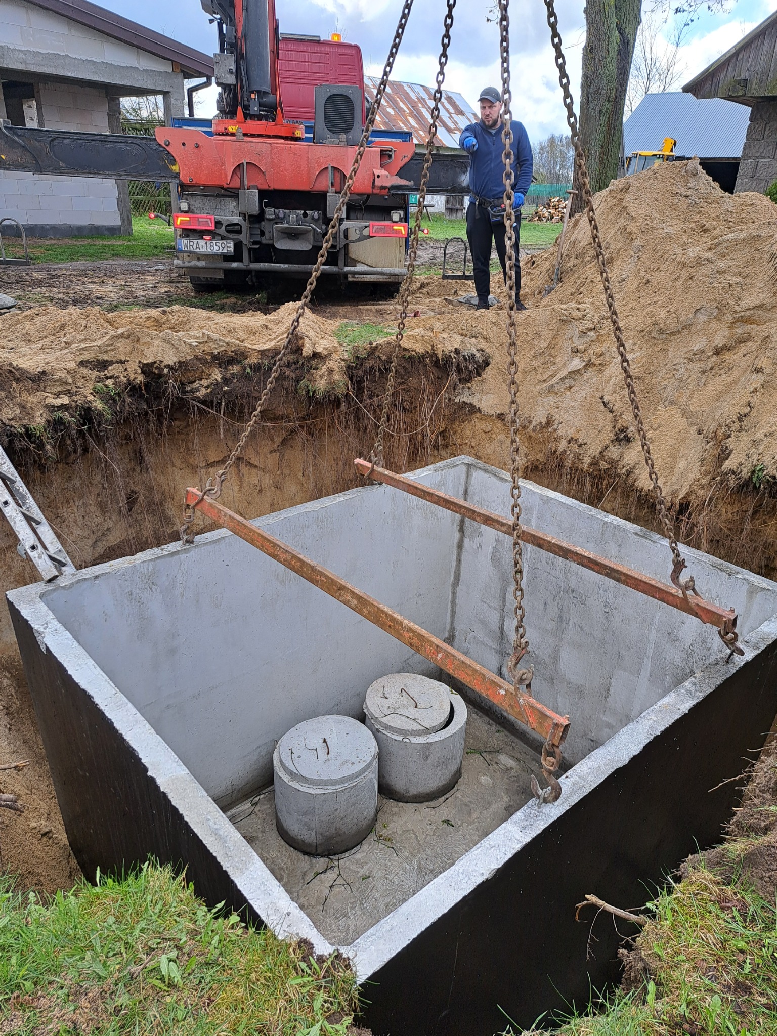 Betonowy zbiornik z dwoma pokrywami opuszczany do wykopu przy użyciu dźwigu samochodowego, pracownik w oddali nadzoruje proces.