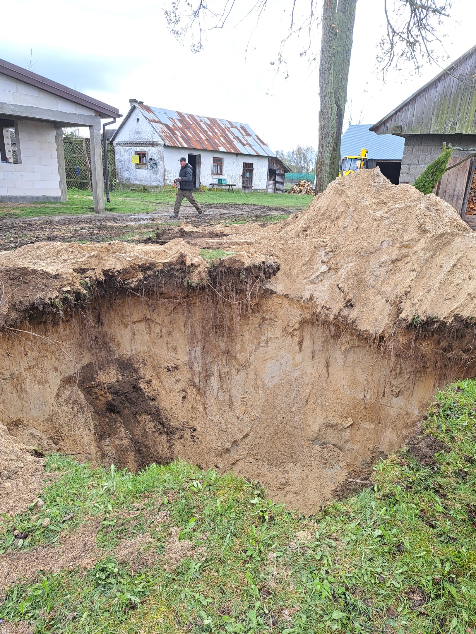 Głęboki wykop w piaszczystej glebie na wiejskim terenie, z pryzmą ziemi obok, w tle budynki gospodarcze i przechodzący mężczyzna z pilarką.