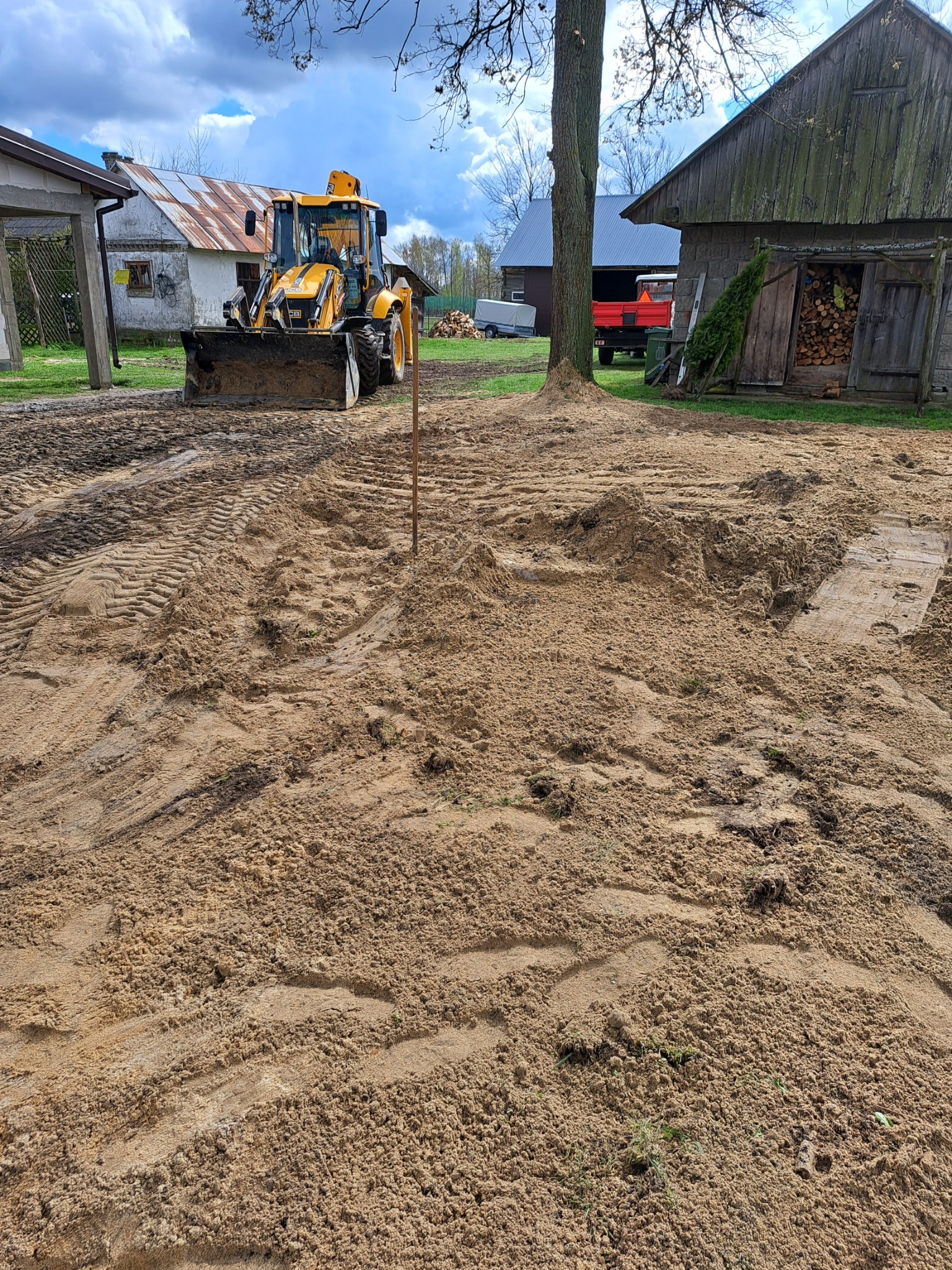 Żółta koparko-ładowarka na piaszczystym terenie z widocznymi śladami opon, w tle budynki gospodarcze i drzewo, pochmurne niebo.