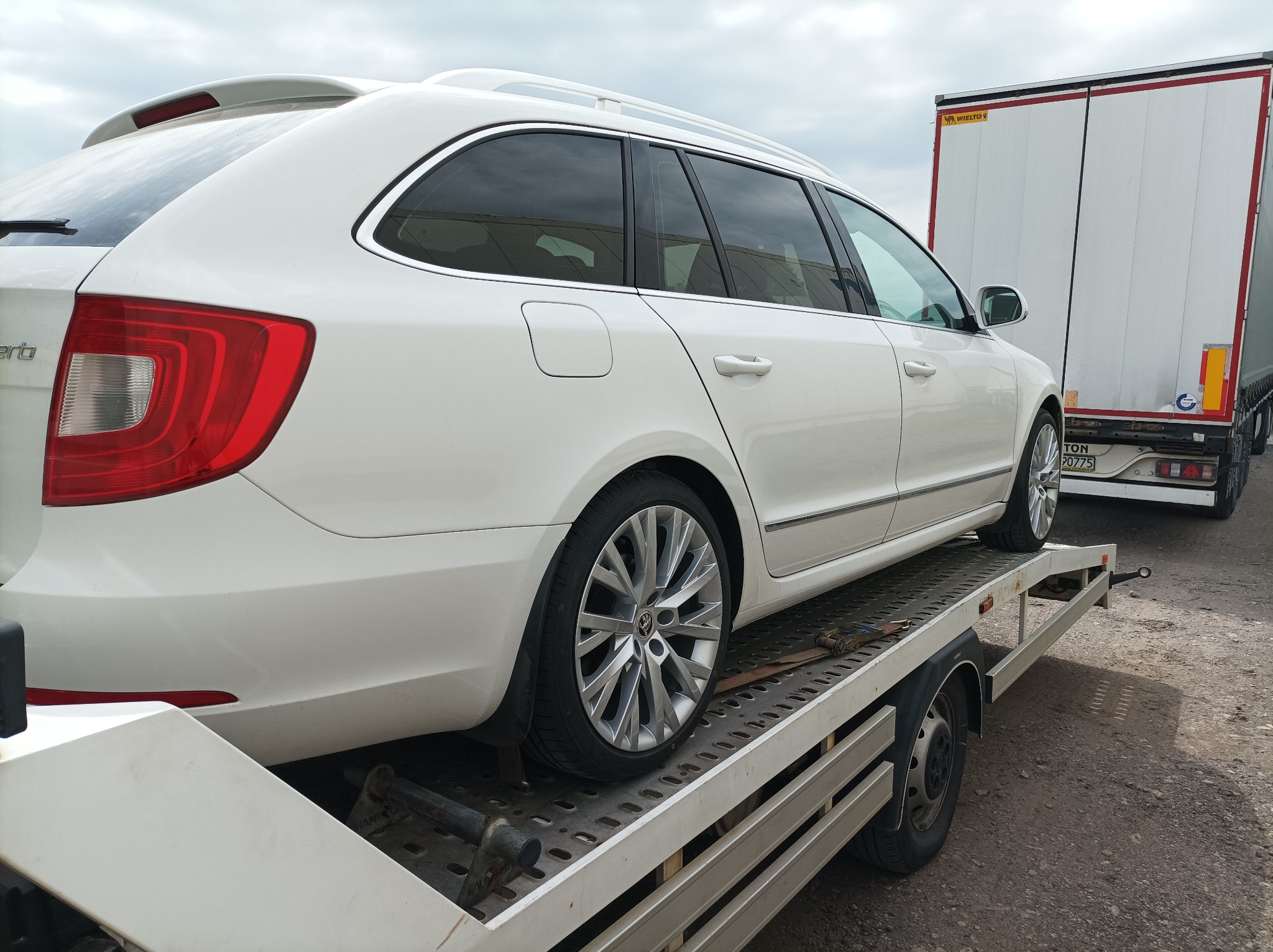 Białe kombi marki Skoda Superb umieszczone na platformie transportowej z widoczną naczepą ciężarową w tle.