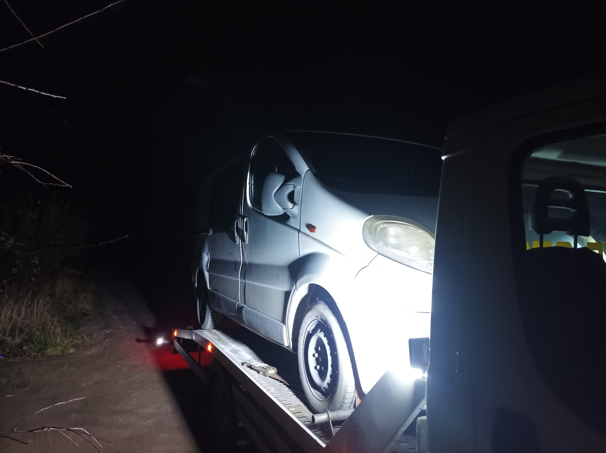 Srebrny bus transportowany na platformie w nocy, oświetlony reflektorem. Widoczna leśna droga w tle.