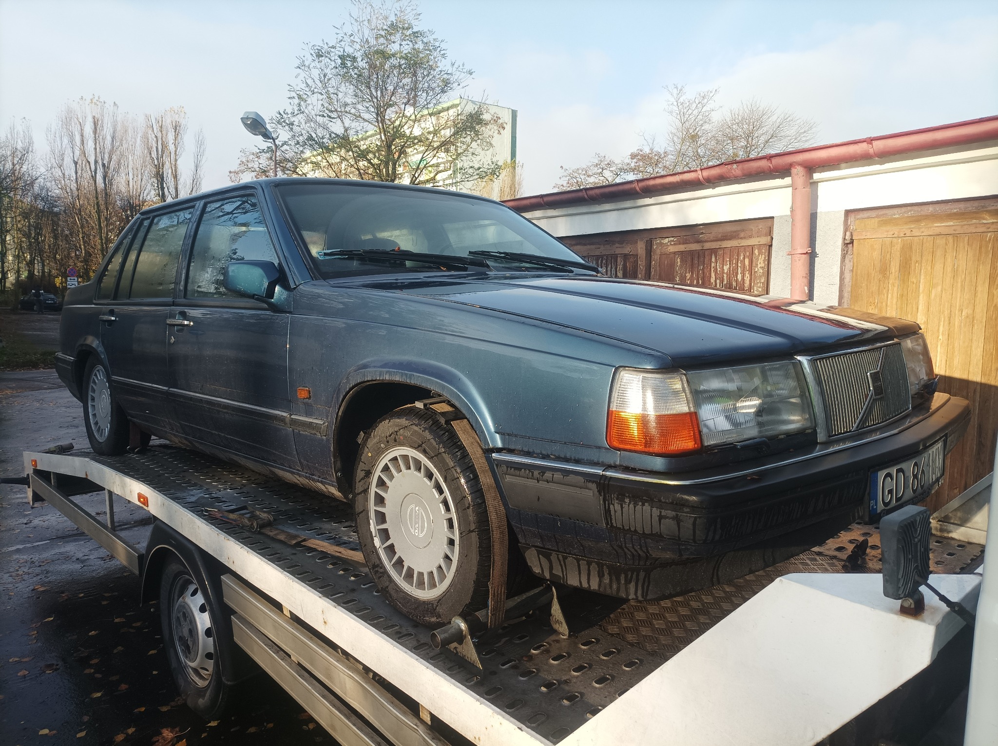 Niebieski sedan Volvo 940 zabezpieczony pasami transportowymi na platformie przyczepy samochodowej z widoczną tablicą rejestracyjną GD 861AN i budynkami w tle.