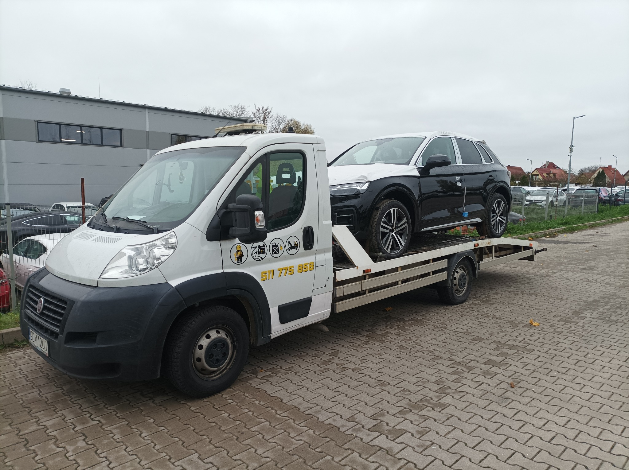Biała platforma transportowa z czarnym Audi Q5 owiniętym folią ochronną na dachu i masce, zaparkowana na brukowanym placu, z budynkiem w tle.