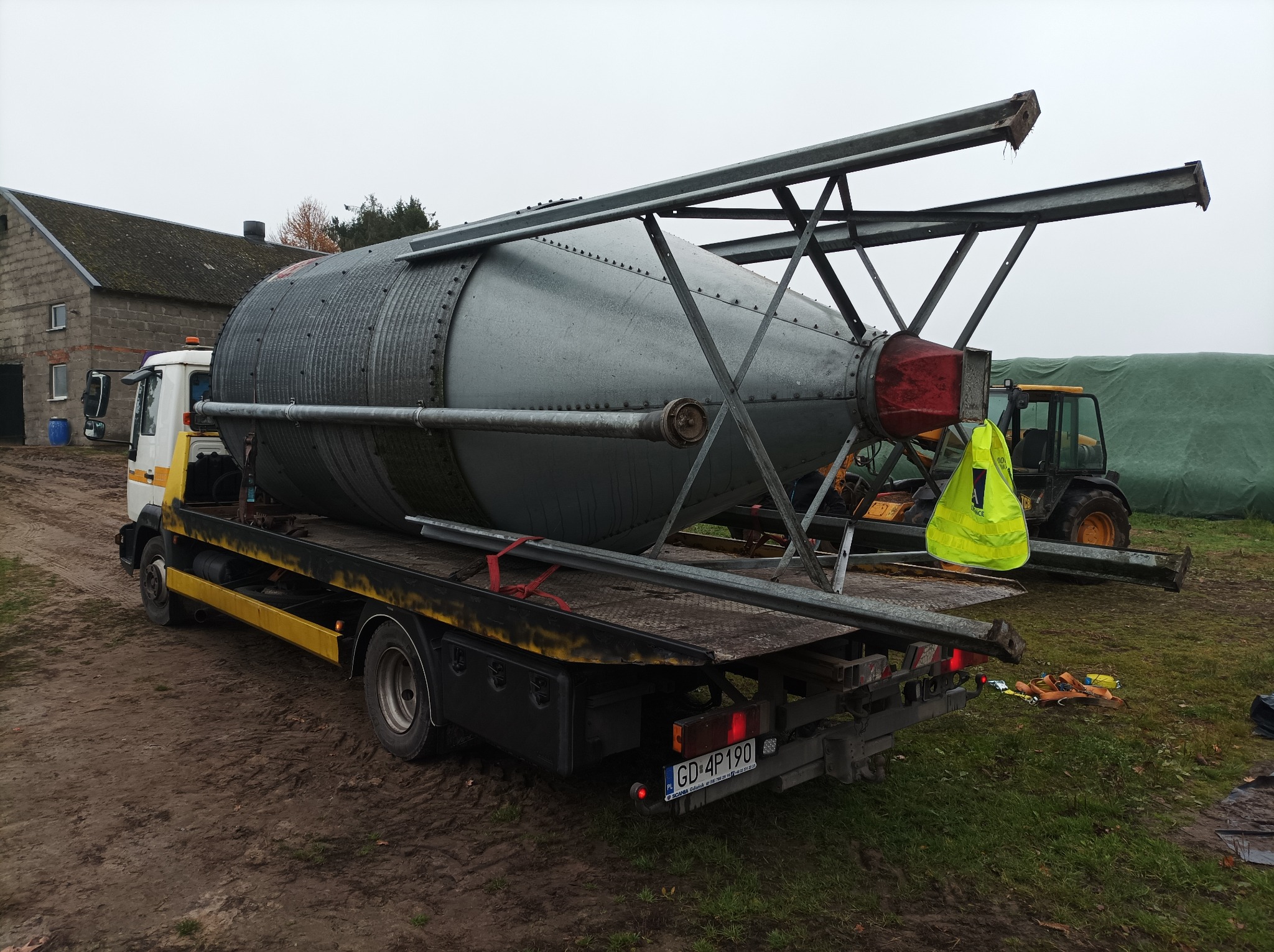 Transport silosu zbożowego na platformie niskopodwoziowej, widoczny żółty kombinezon roboczy i traktor w tle, teren błotnisty.