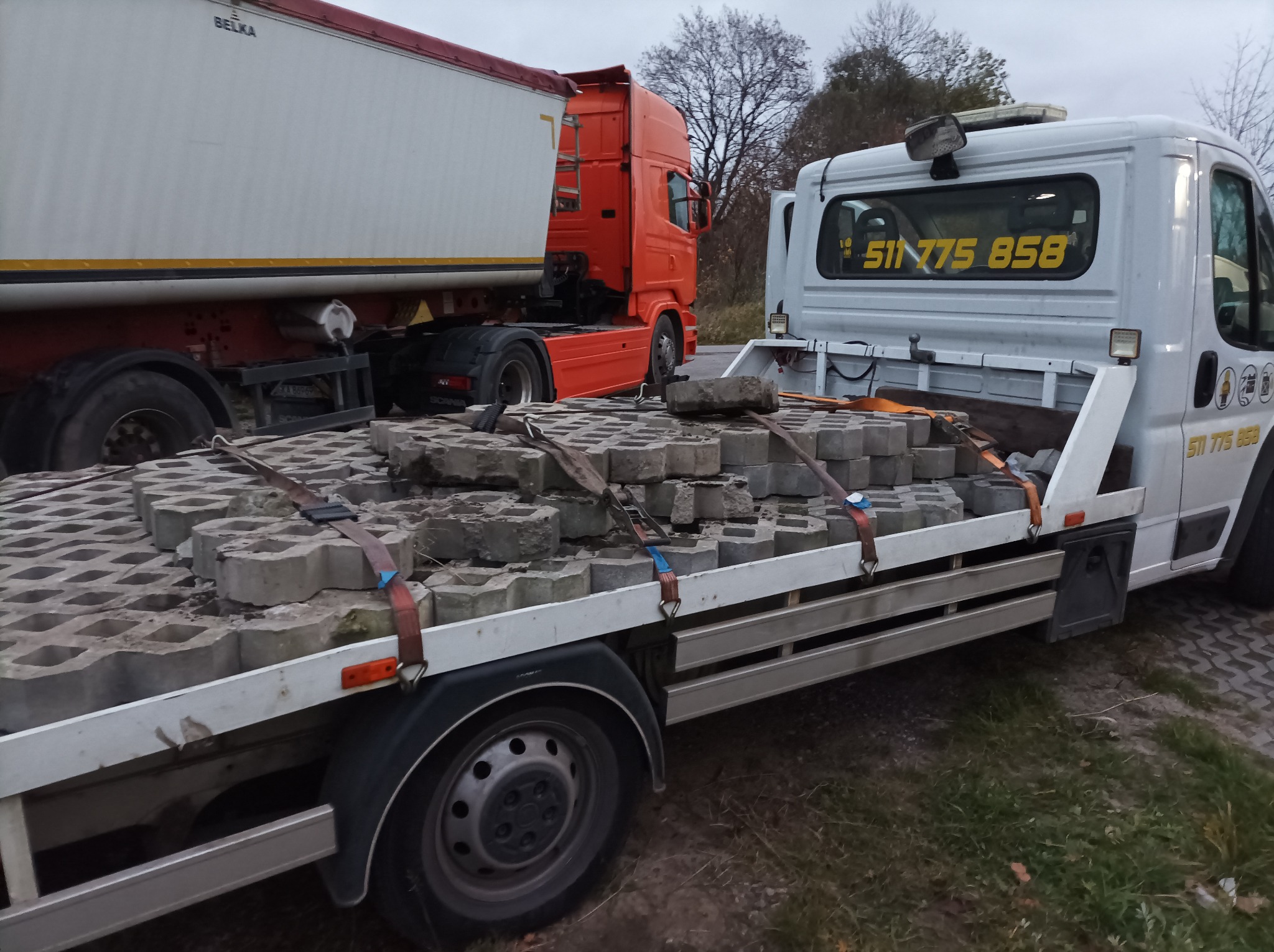 Transport kostki ażurowej na platformie pojazdu z numerem telefonu, w tle pomarańczowa ciężarówka z białą naczepą.