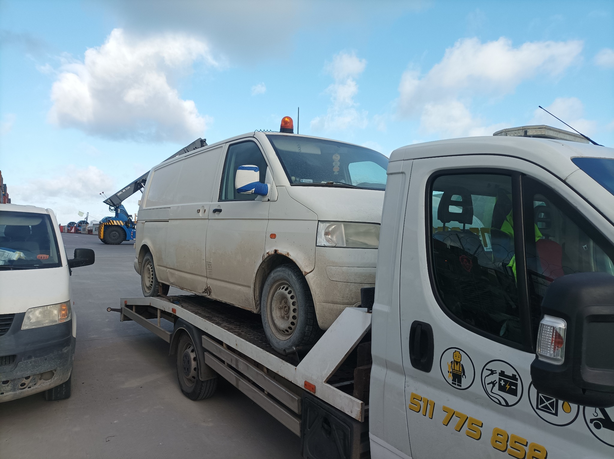 Biały, zabrudzony furgon z pomarańczowym światłem ostrzegawczym na dachu, przewożony na platformie transportowej; widoczny fragment drugiego pojazdu holowniczego z naklejkami informacyjnymi...