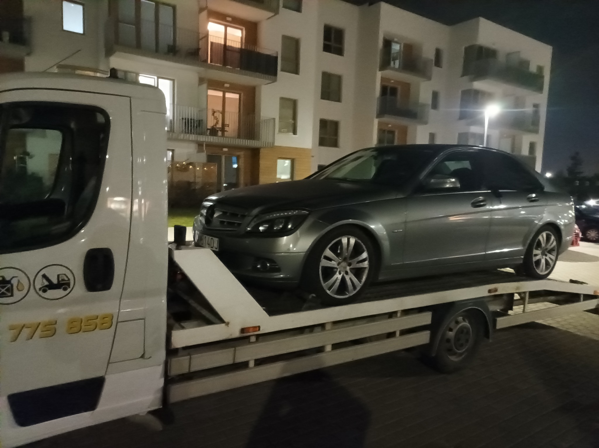 Srebrny samochód osobowy Mercedes-Benz na białej platformie transportowej, widok nocny na tle oświetlonego bloku mieszkalnego.